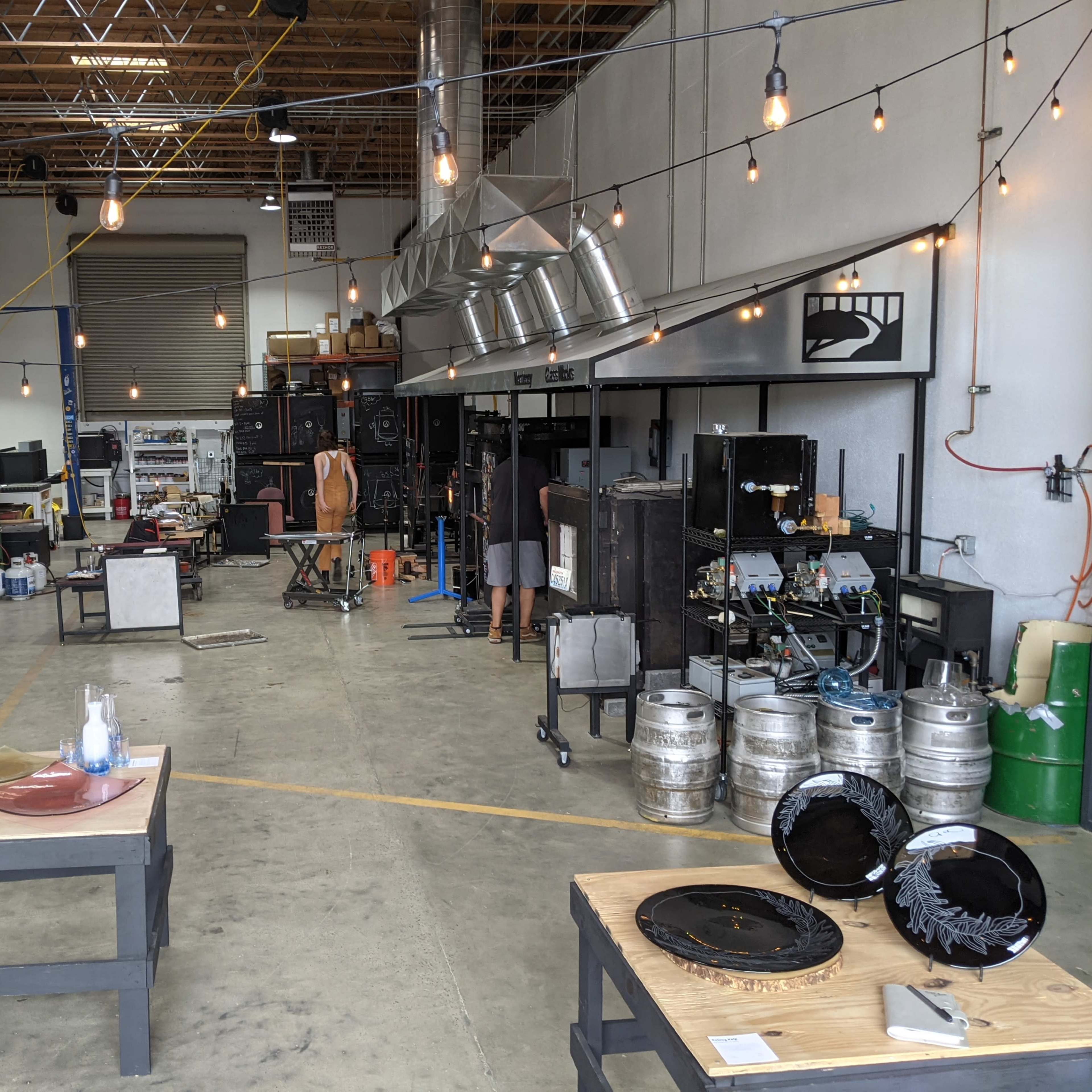 A spacious workshop with various equipment and tools, illuminated by hanging light bulbs, featuring glassware and metal trays on wooden tables.