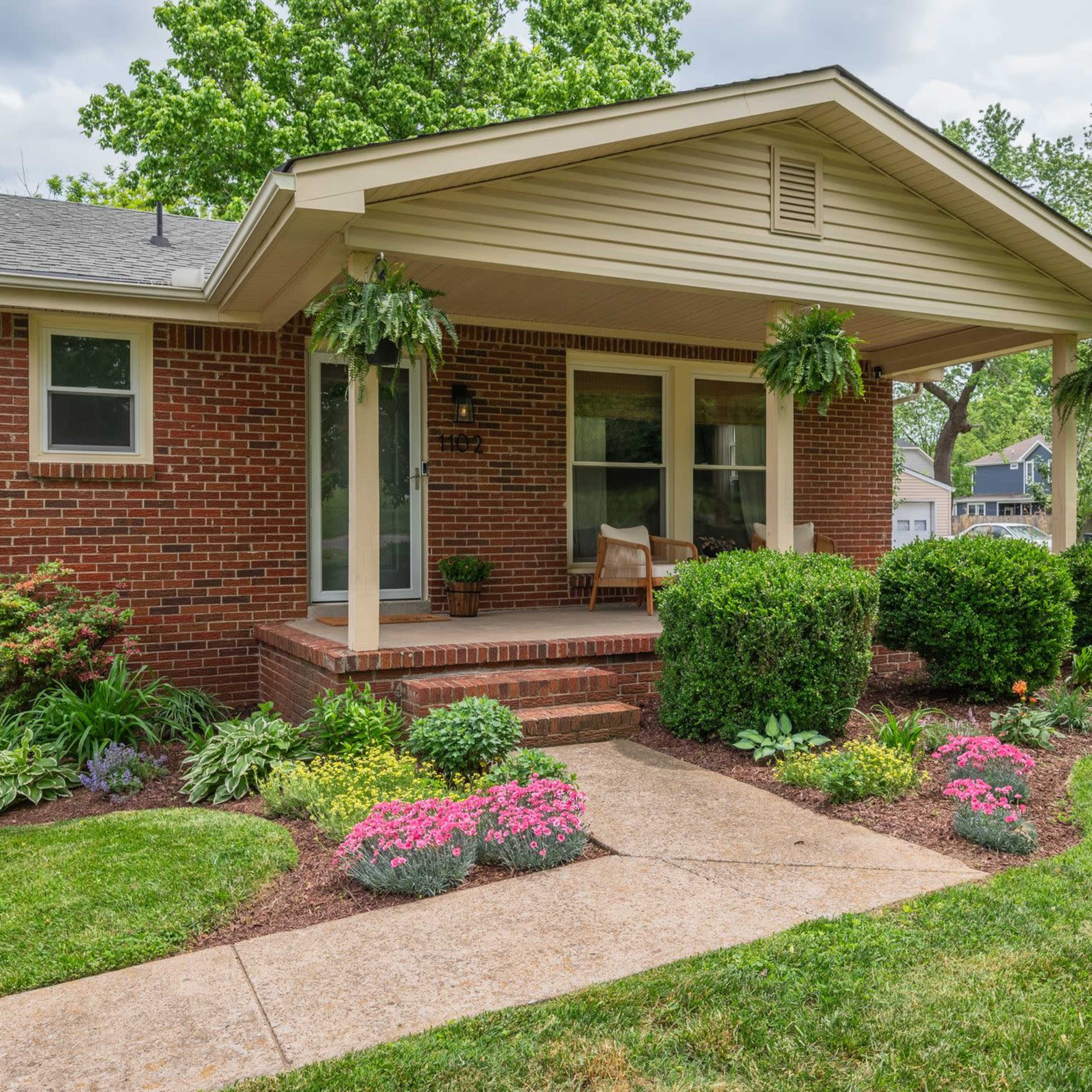 A brick house with a front porch, flanked by decorative plants and flower beds along a walkway.