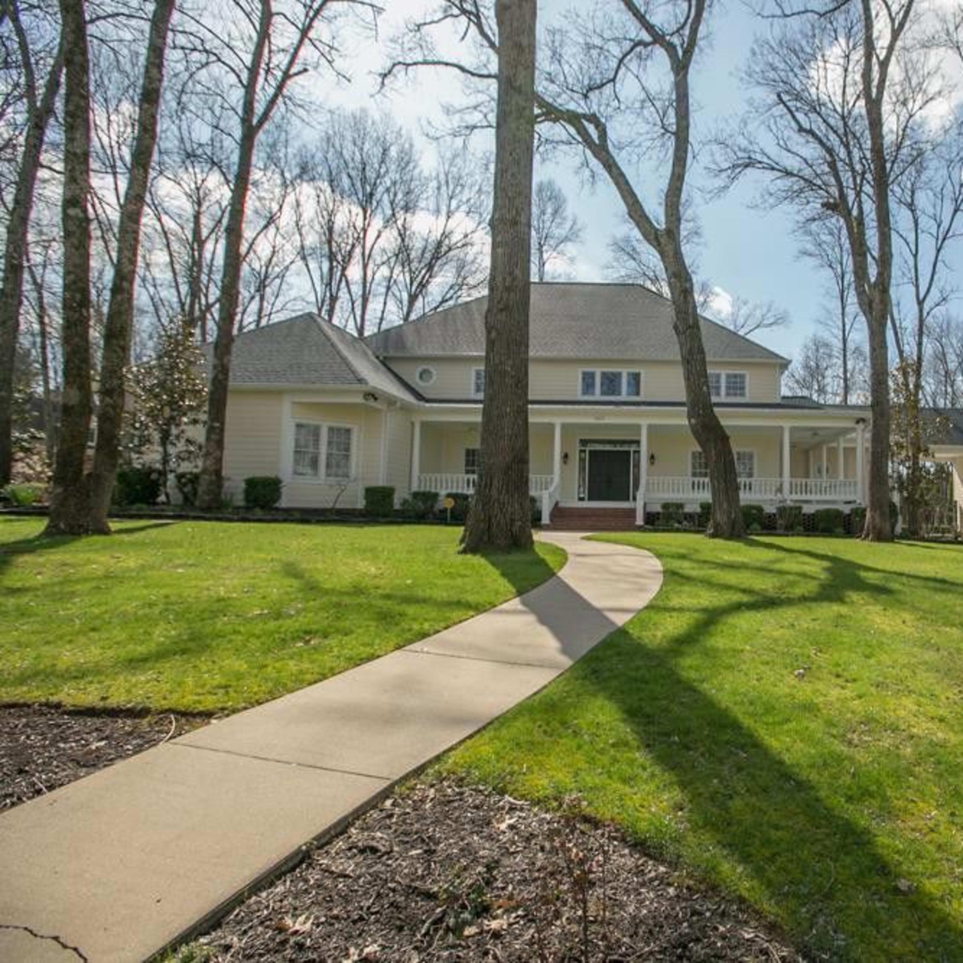 A spacious two-story house is surrounded by tall trees and a manicured lawn, with a winding walkway leading to the entrance.