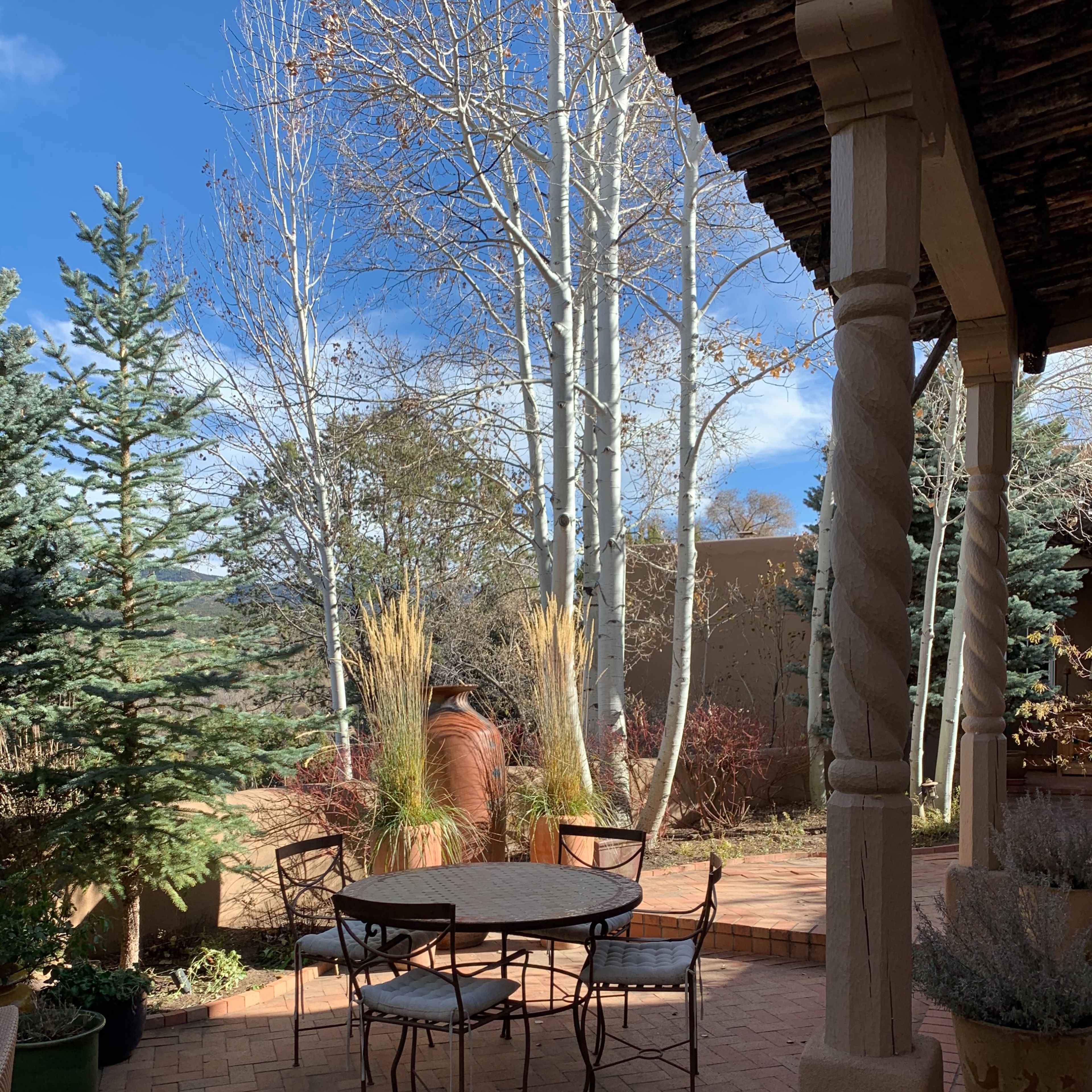A small round metal table with four chairs is set on a brick patio surrounded by trees and shrubs.