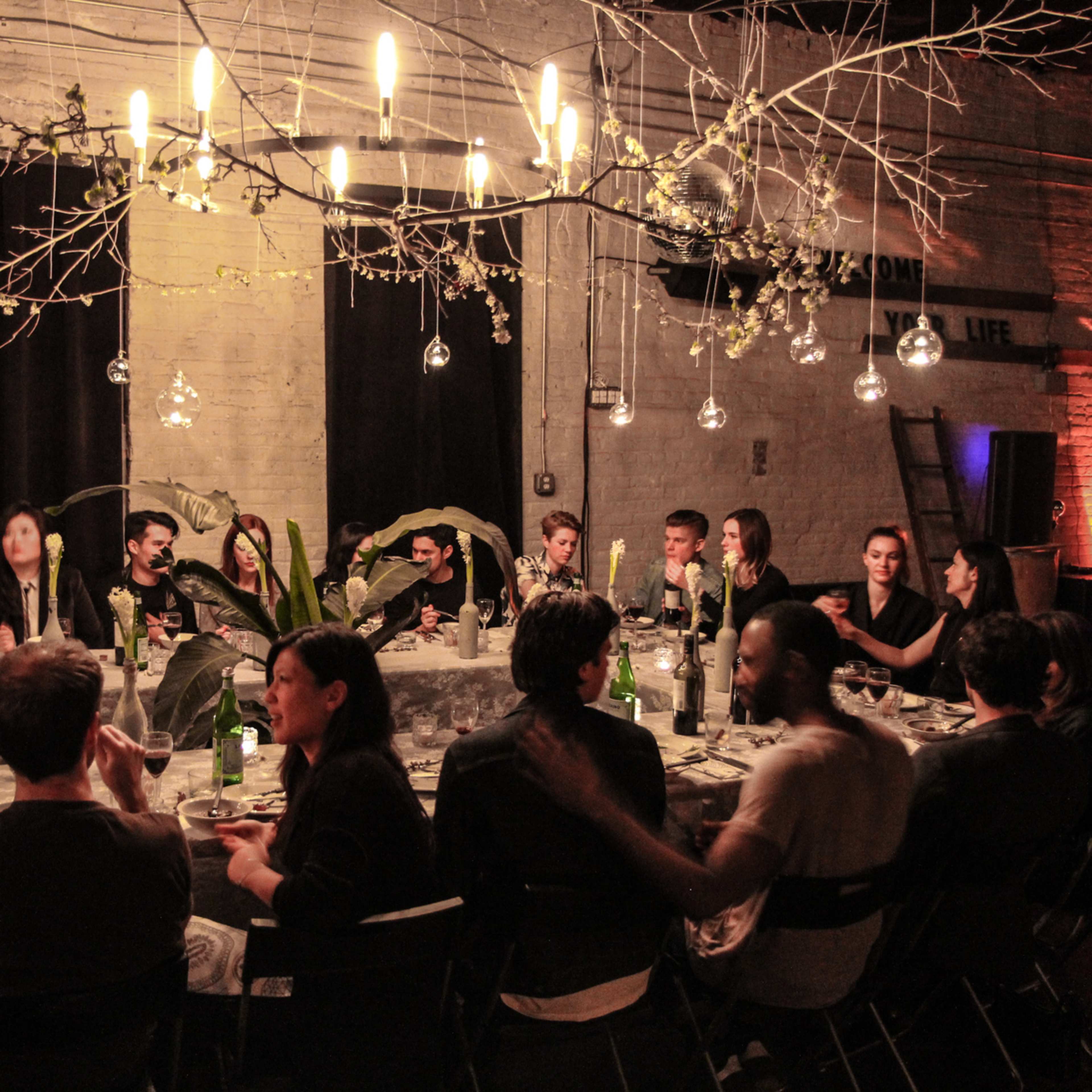 A spacious room with wooden floors, large windows, a grand piano, and various seating areas adorned with decorative plants and rugs.