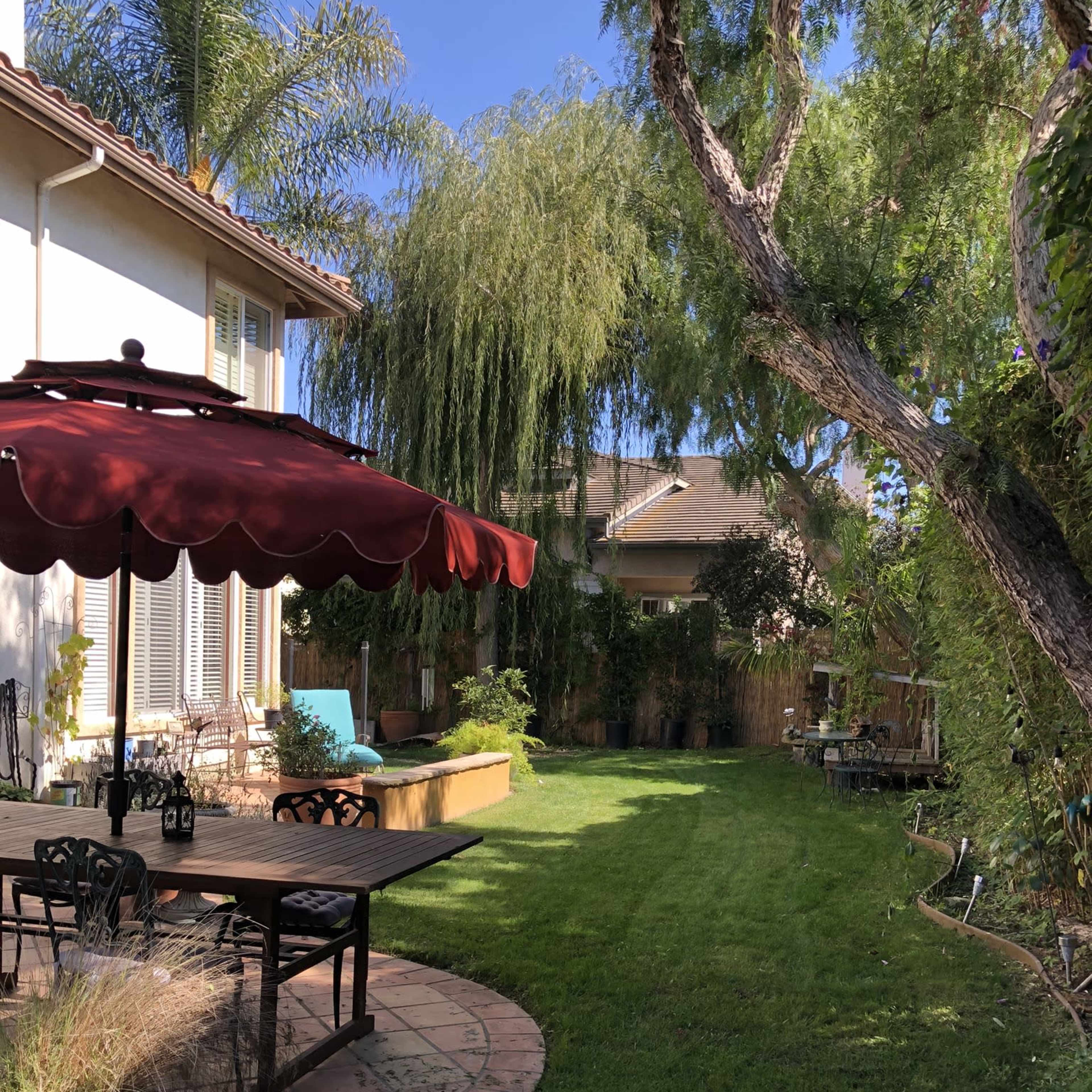 A backyard features a patio with a shaded table and chairs, surrounded by well-kept grass and lush greenery.