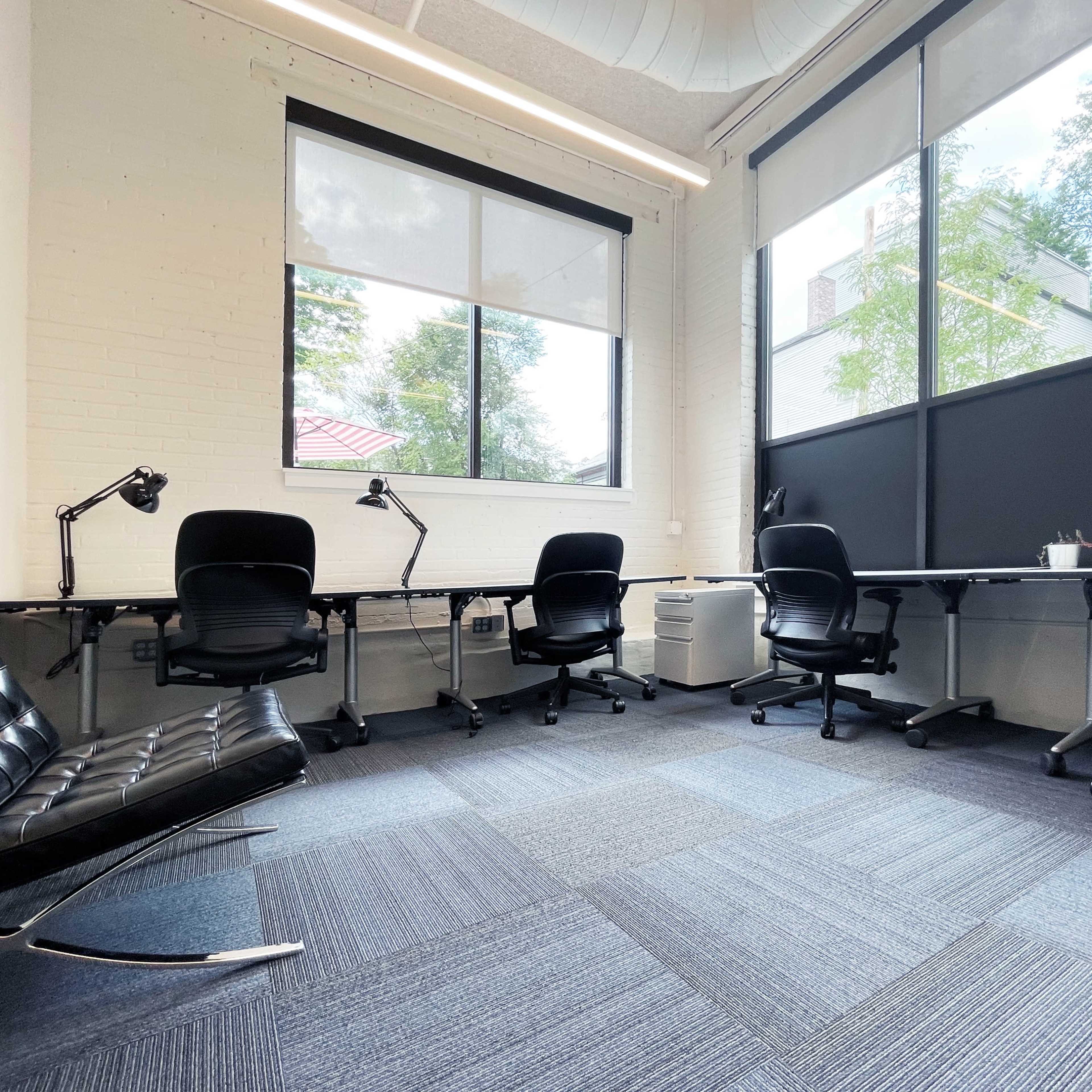 A modern office space with four black chairs, a black lounge chair, and desks arranged near large windows that allow natural light to enter.