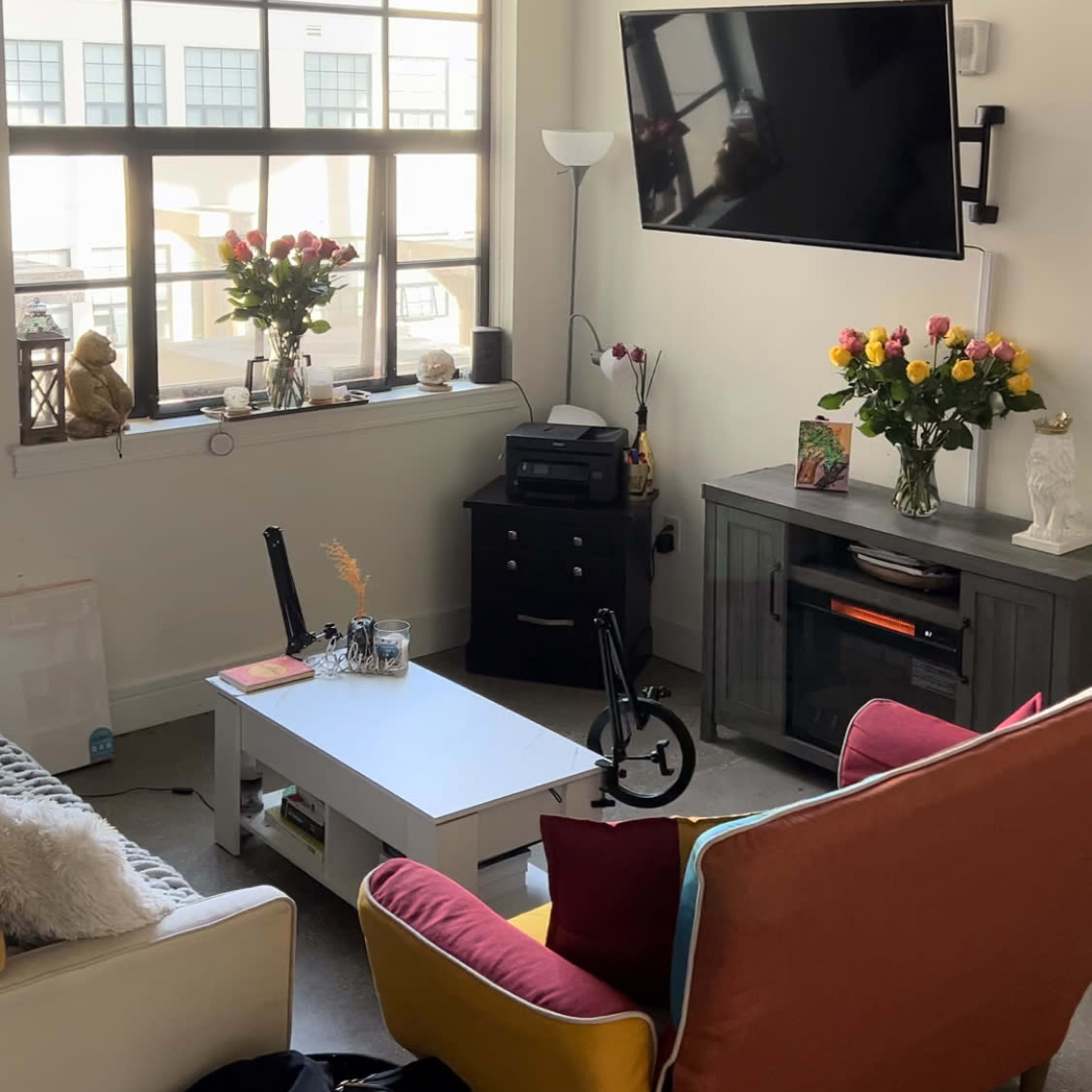 A modern living room with a large window, a wall-mounted TV, a coffee table, and colorful seating arrangements, along with flower arrangements on a side table.