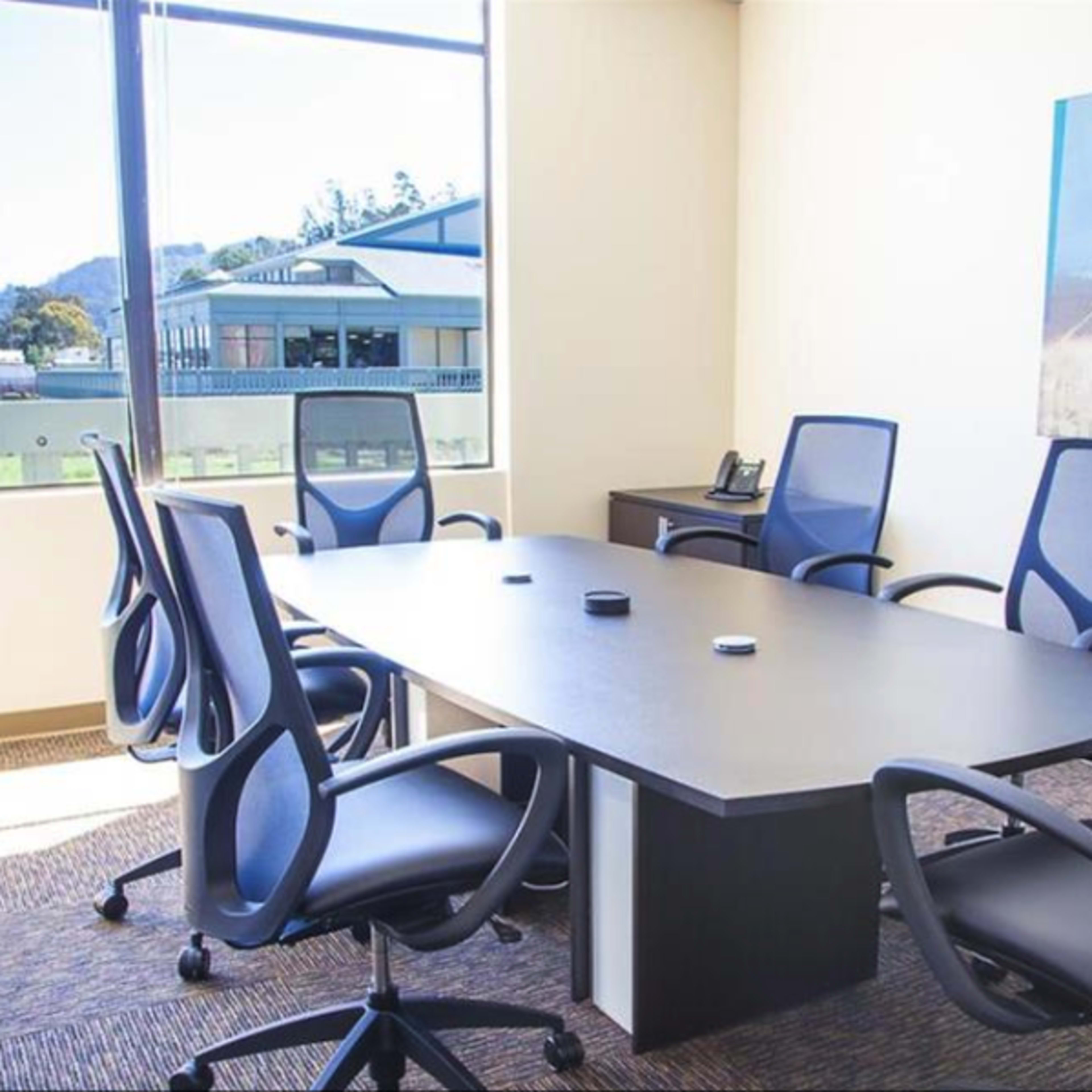 A conference room features a large table surrounded by six mesh office chairs, with a window providing a view of the outside.