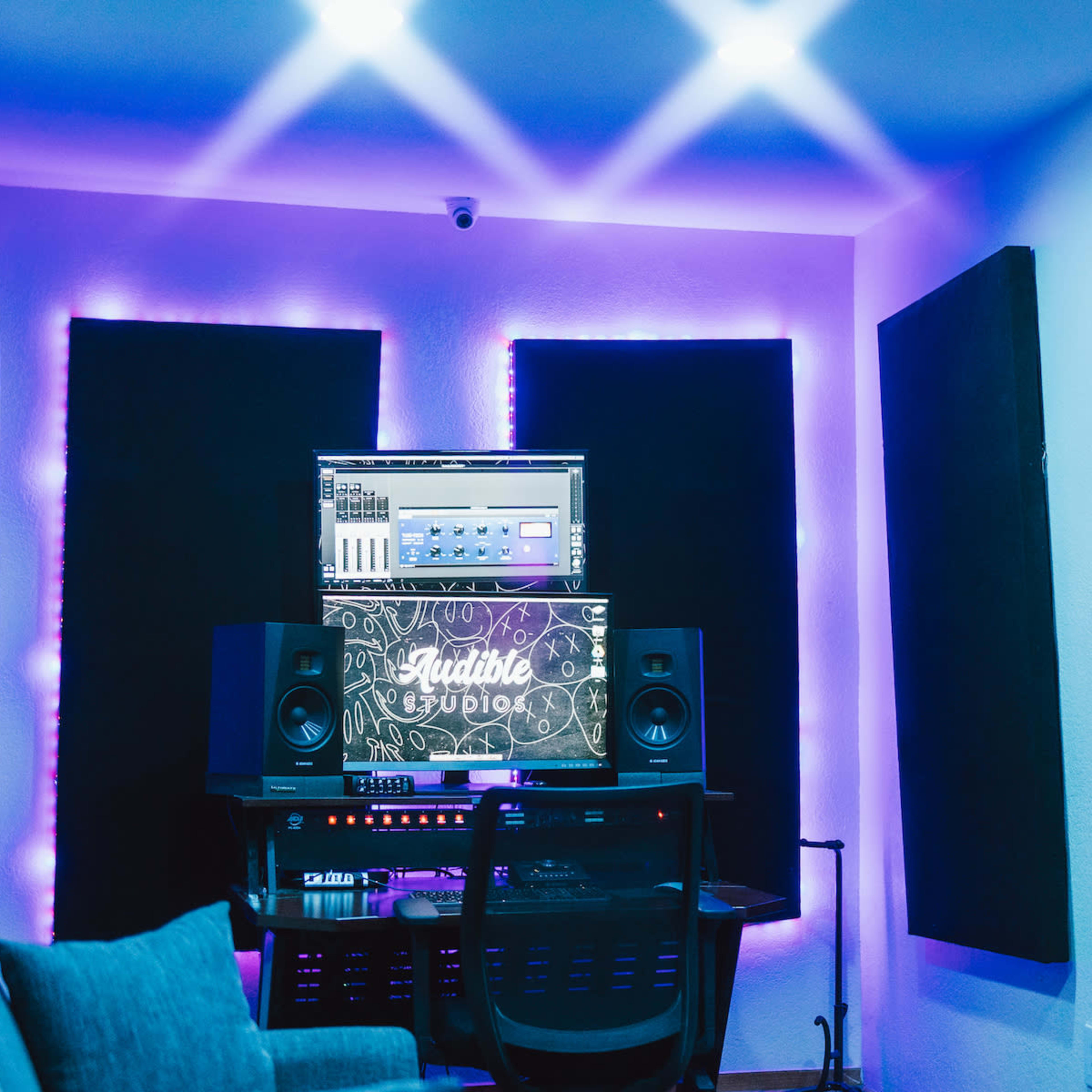 A music recording studio features dark soundproof panels and a computer screen with audio software, illuminated by purple LED lights.