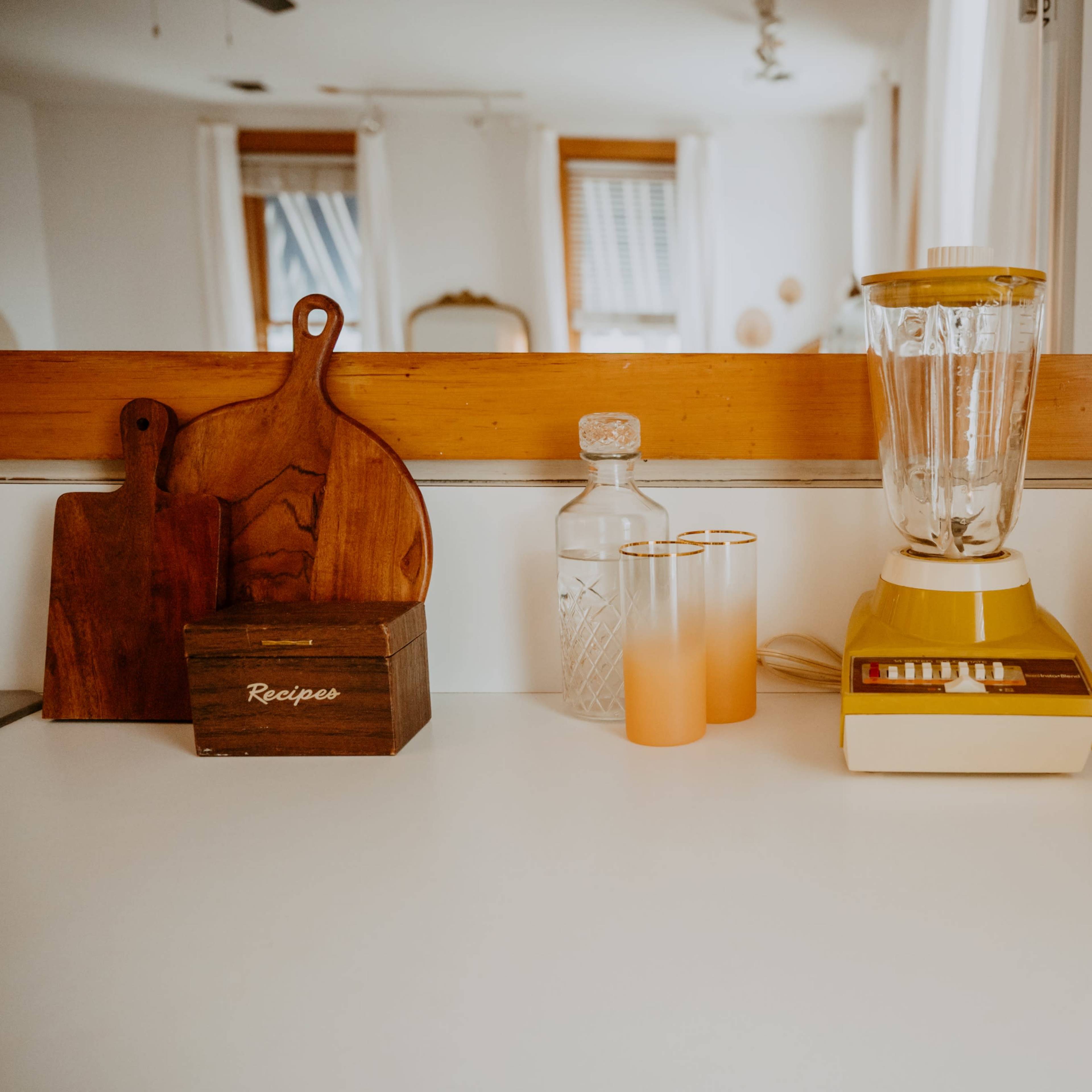 A kitchen countertop with a blender, colored glasses, a glass bottle, wooden cutting boards, and a retro radio.