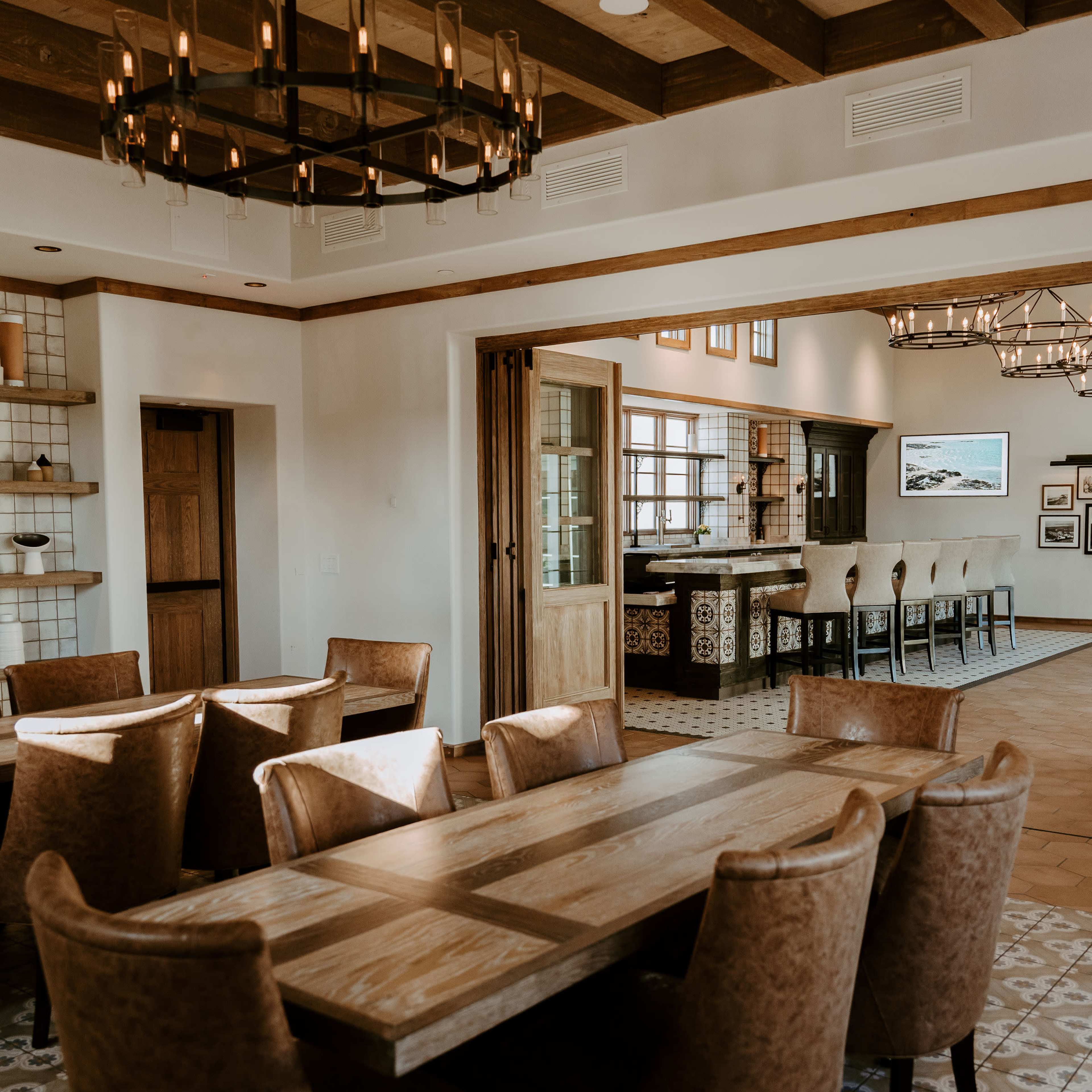 A dining area featuring a long wooden table surrounded by upholstered chairs, with a view of an adjacent kitchen area in a well-lit, modern interior.