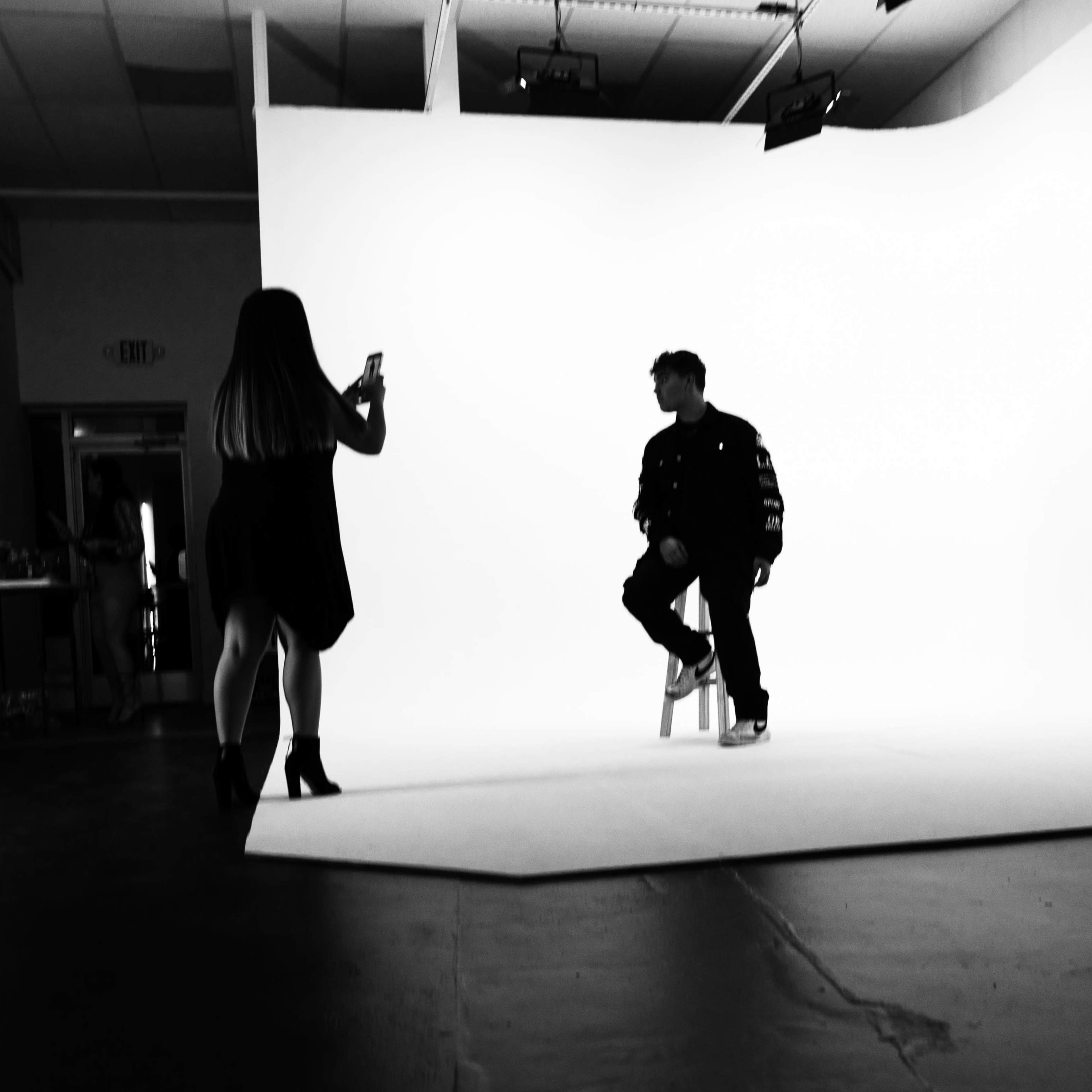 A person in a black jacket sits on a stool against a white backdrop while another person takes a photo of them.