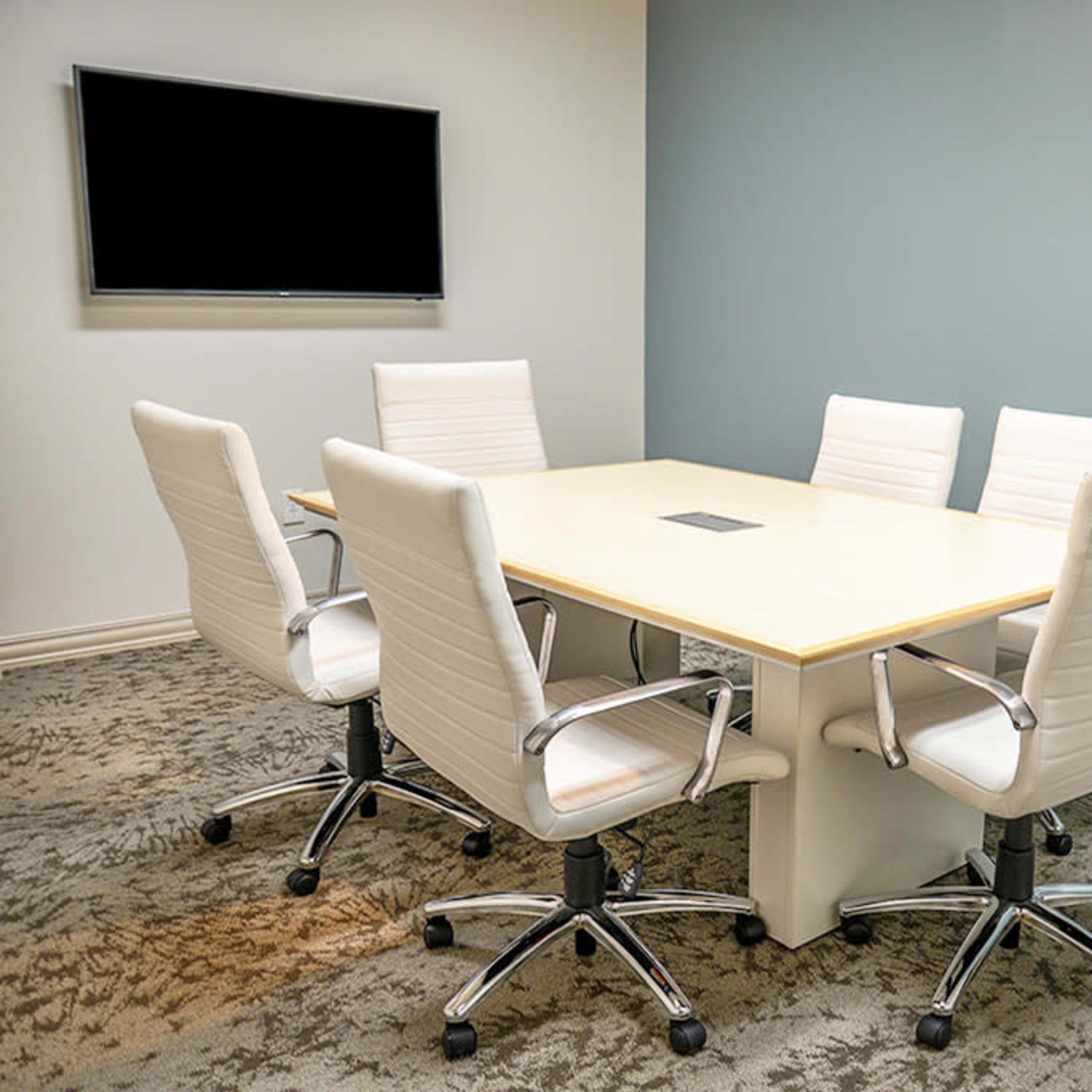 A conference room features a rectangular table surrounded by five white chairs and a wall-mounted screen.