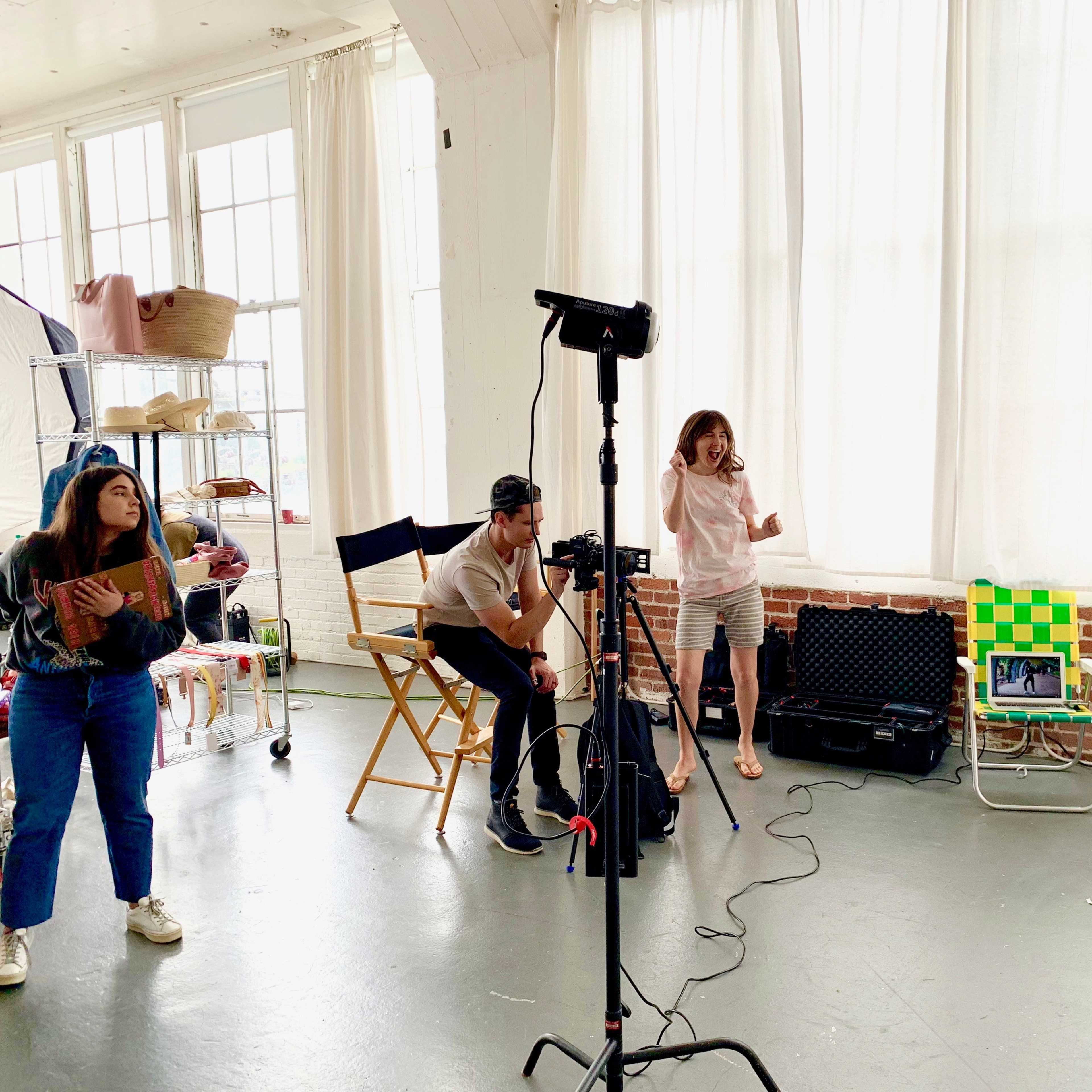 A group of three individuals is working in a bright studio filled with fabric racks and large windows while preparing for a photoshoot.