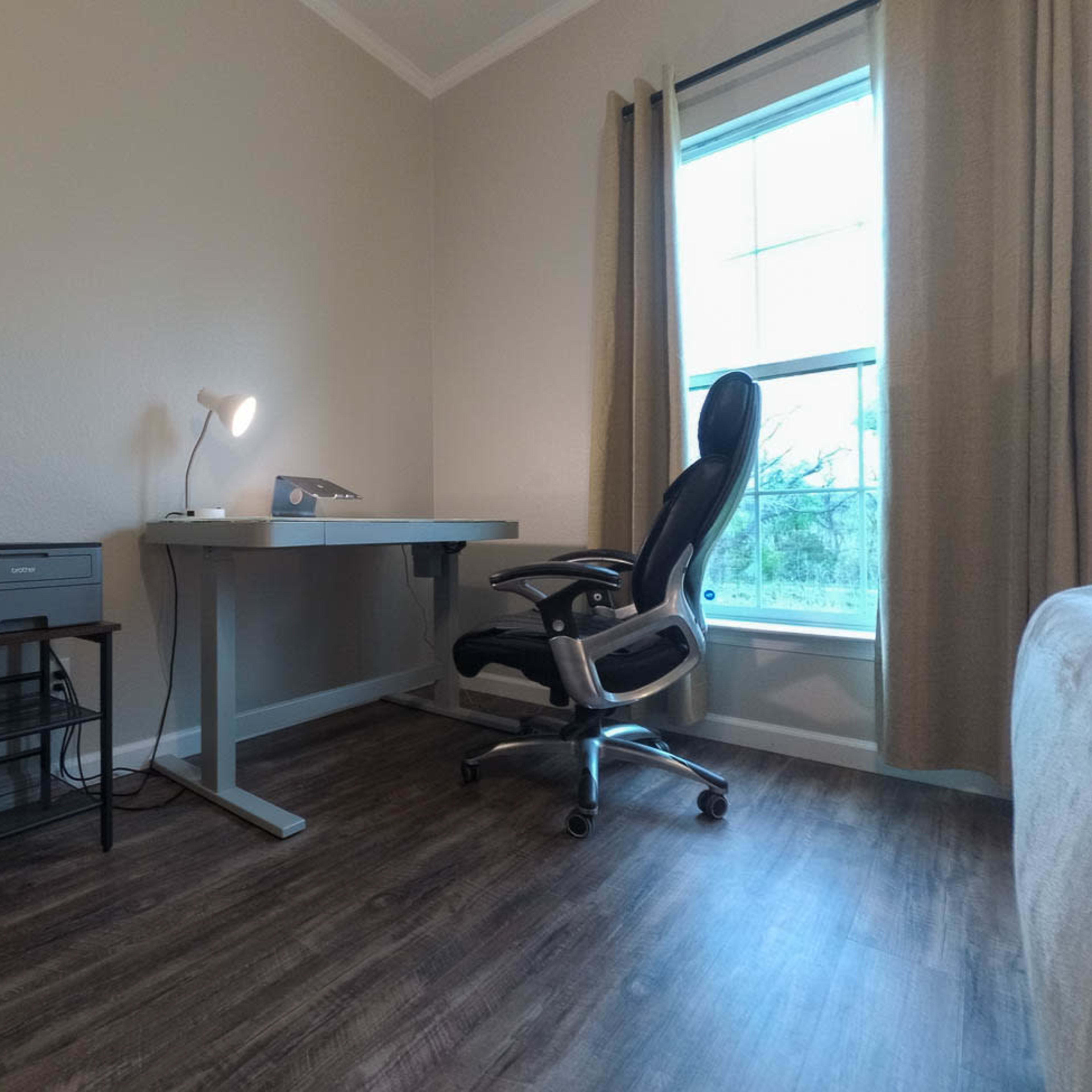 A minimalist home office setup featuring a desk, an office chair, a printer, and a window with curtains.