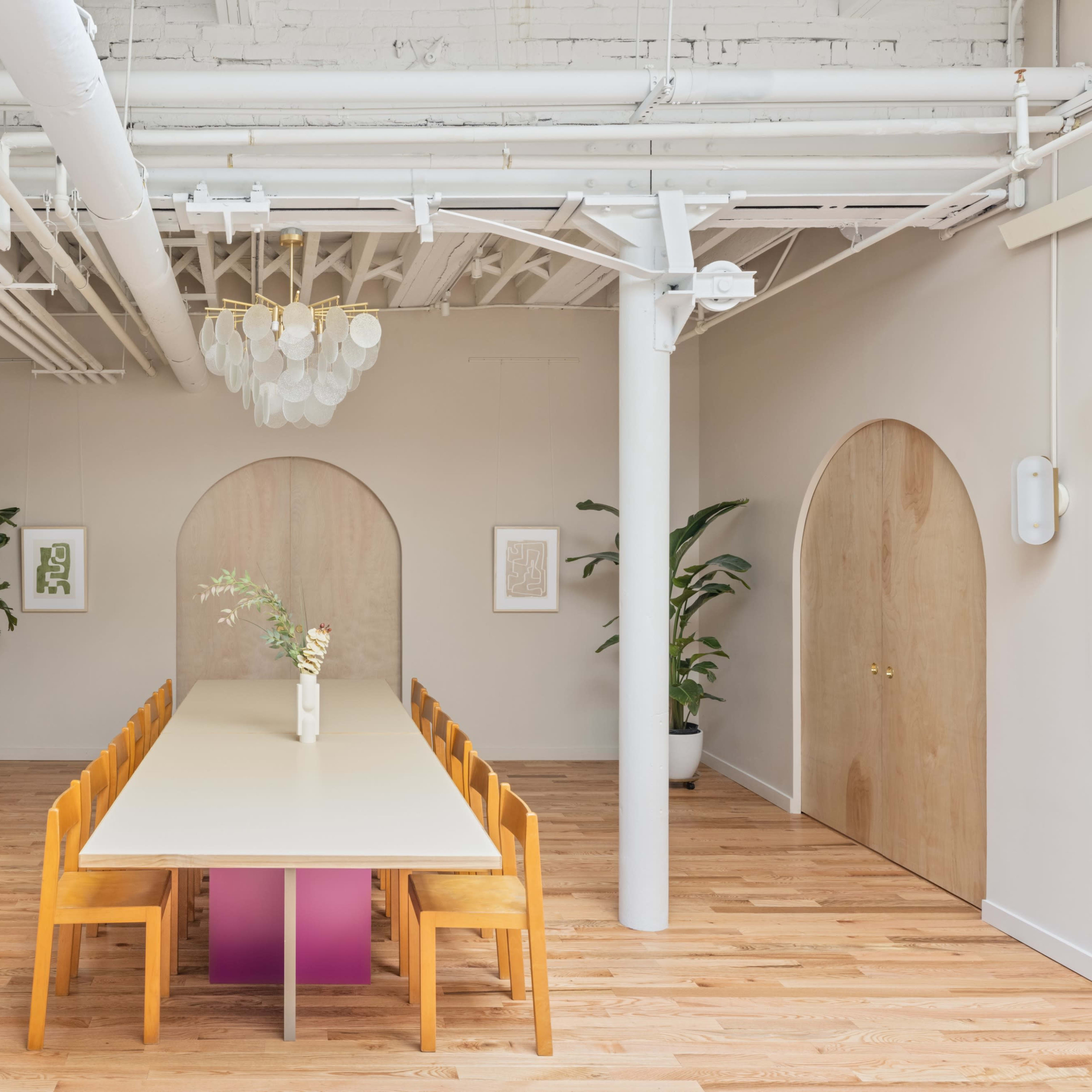 A long table surrounded by wooden chairs is positioned in a bright, modern room with arched doorways and potted plants.