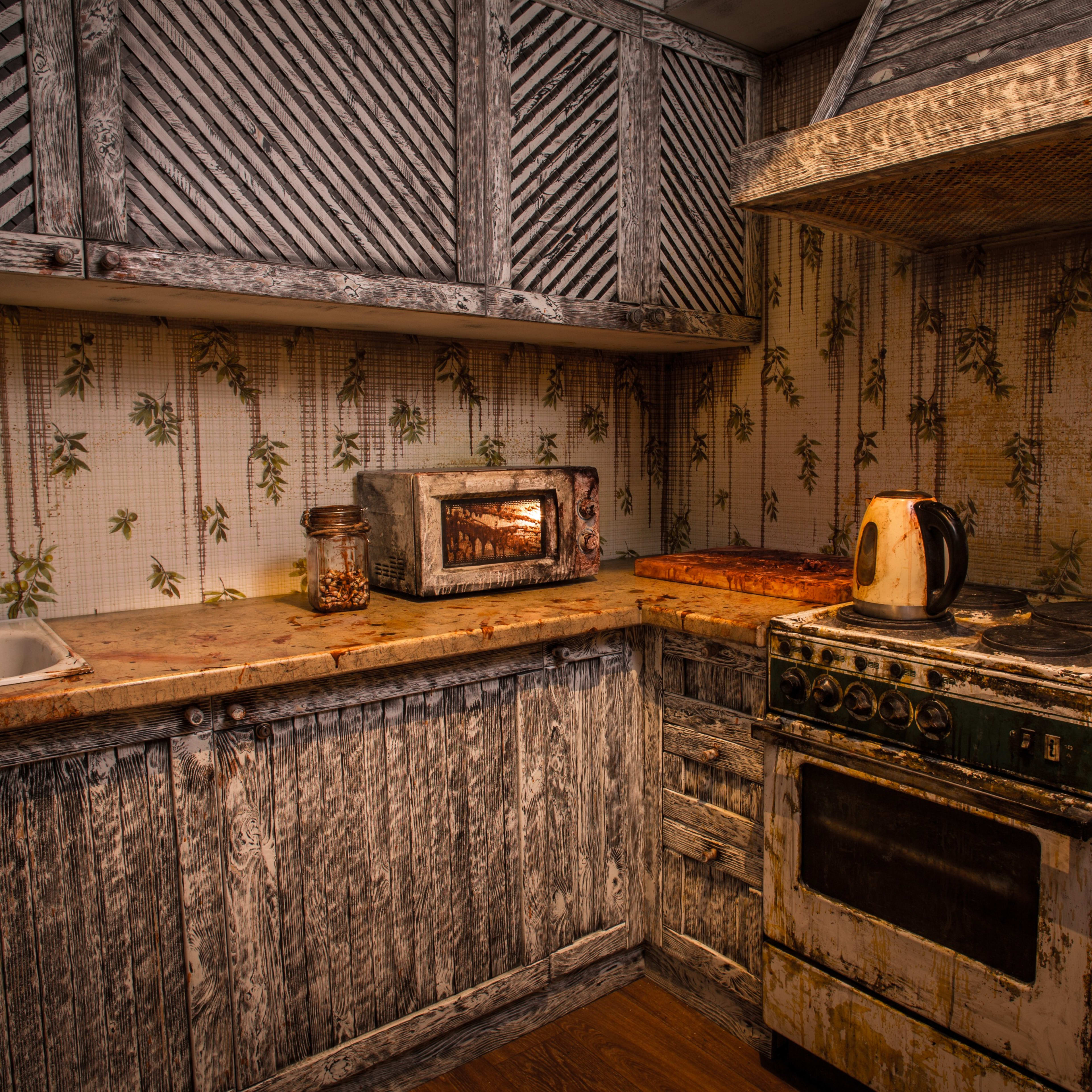 A weathered kitchen with a rustic wooden cabinet, an old stove, a microwave, and a kettle on a stained countertop.