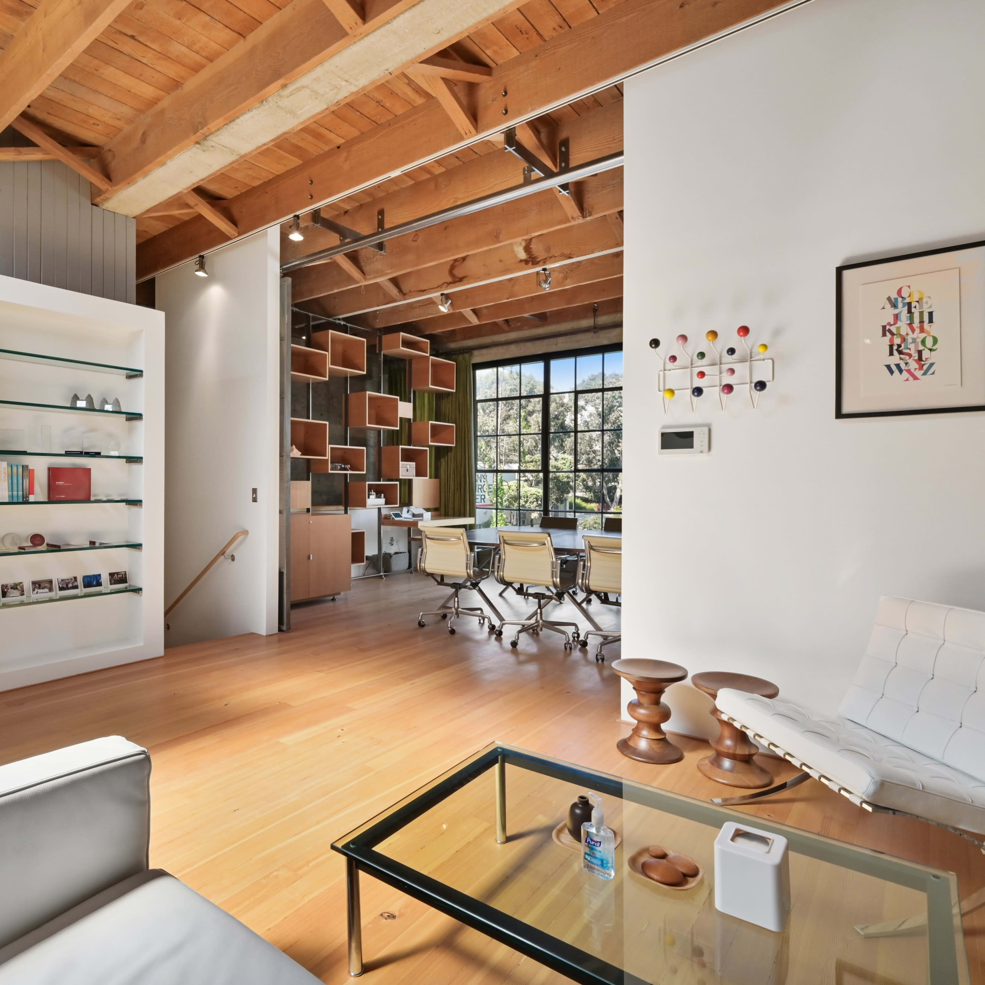 A modern office space with wooden beams, a glass coffee table, a white sofa, and a workstation surrounded by bookshelves.