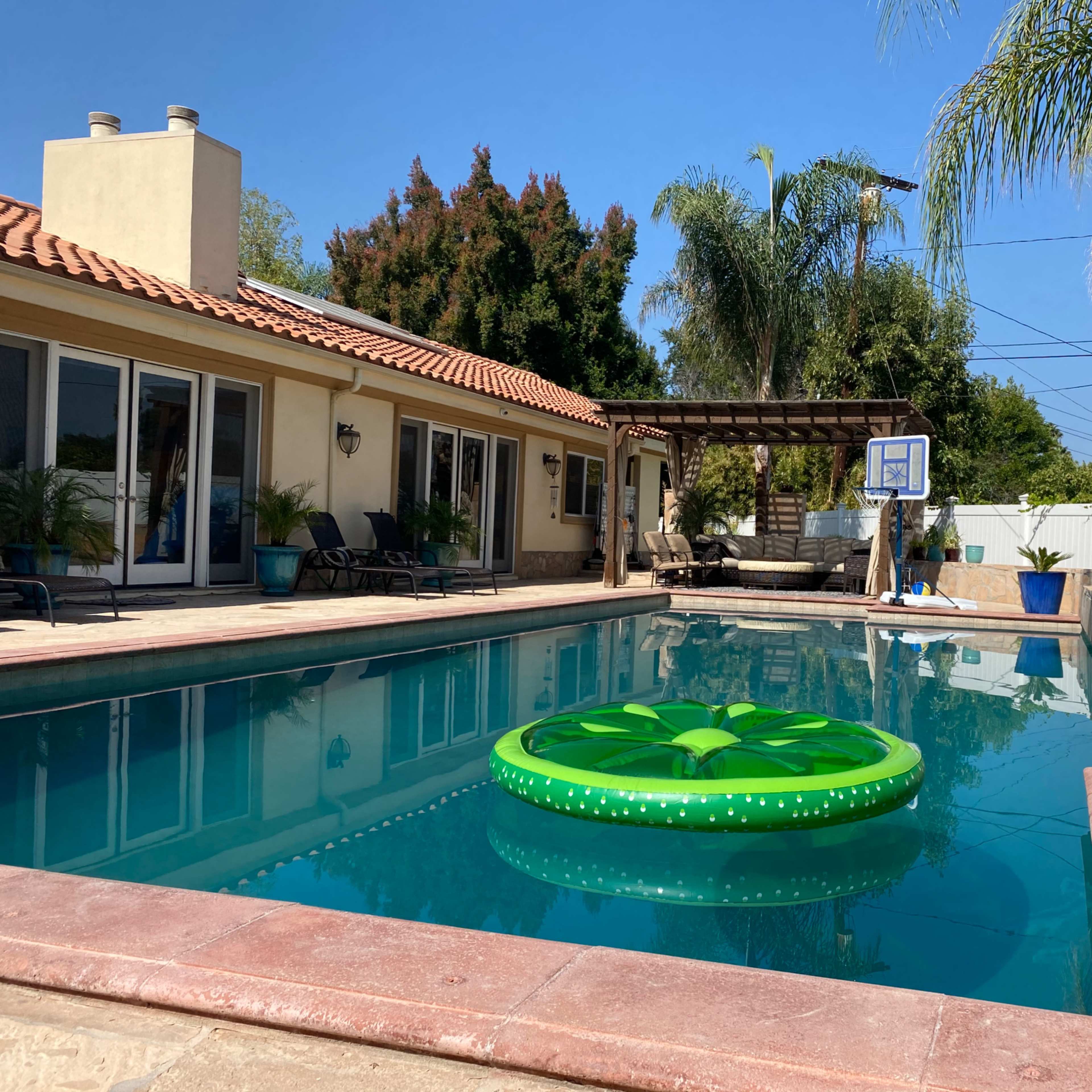 A swimming pool with a green inflatable raft is surrounded by a patio and tropical plants under a clear blue sky.