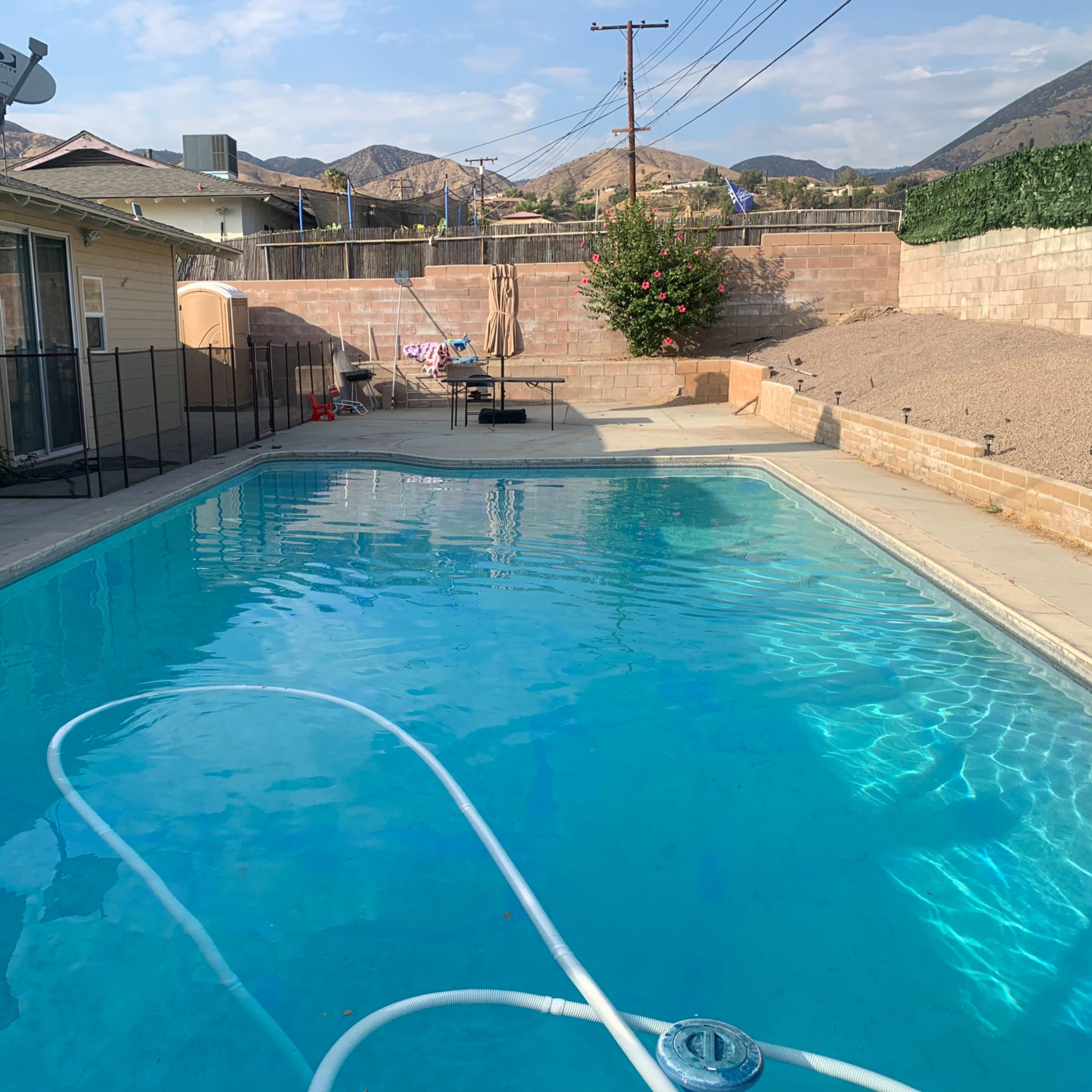 The scene shows a clear swimming pool surrounded by a fenced area, with mountains in the background and patio furniture nearby.
