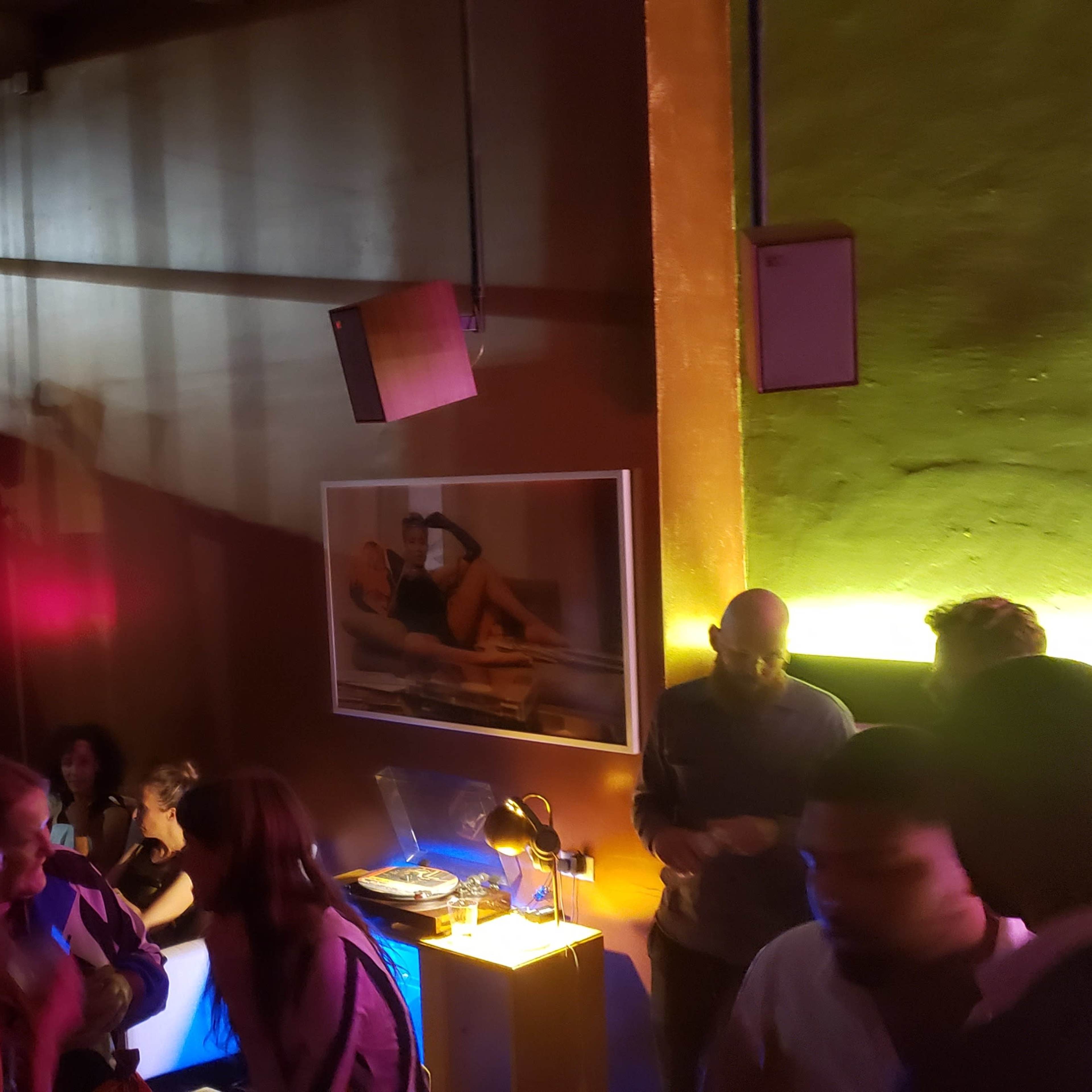The scene depicts a dimly lit indoor space with people gathered around a bar area, colorful lighting, and a framed photo on the wall.