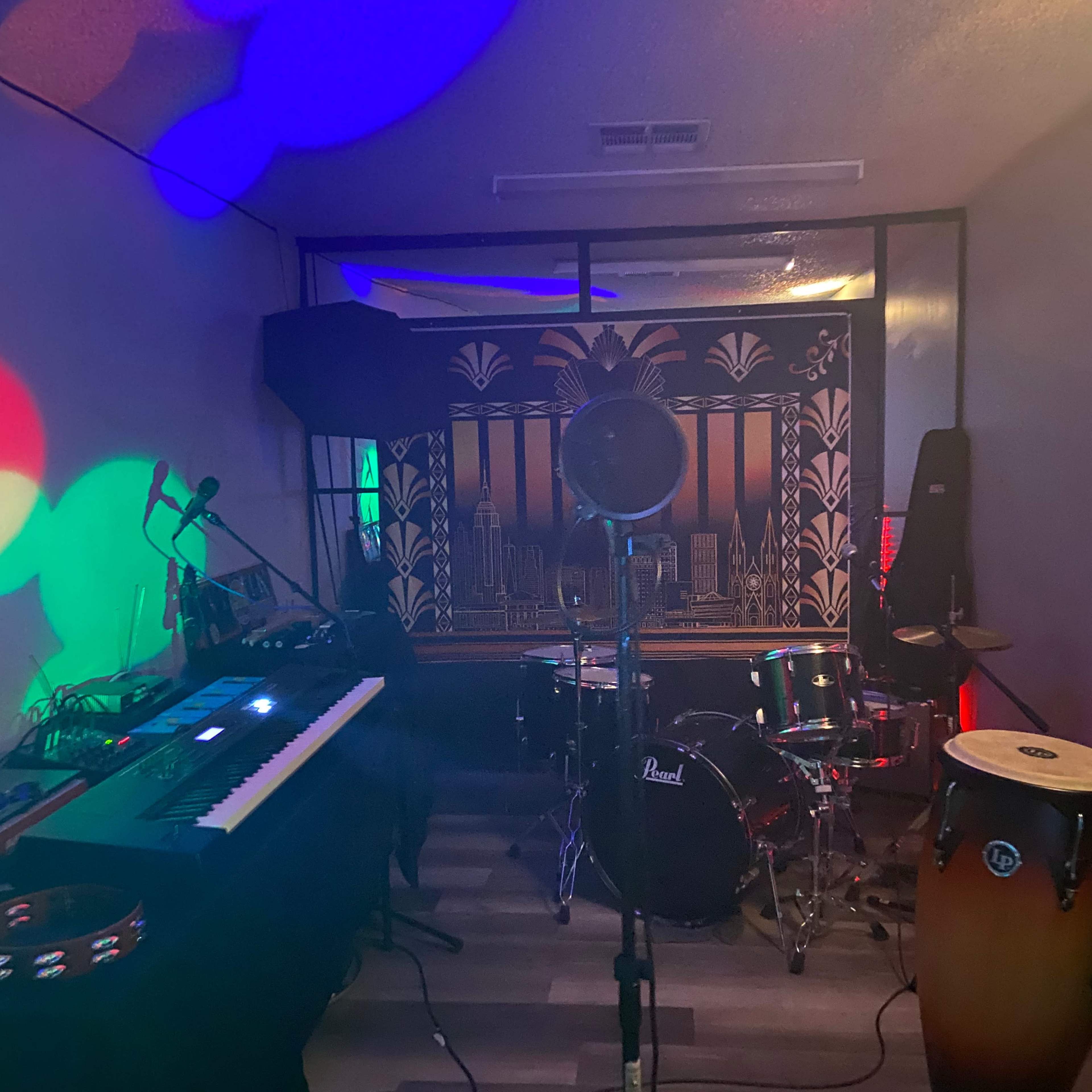 A music studio setup featuring a keyboard, drums, conga drums, and colorful lighting effects on the walls.