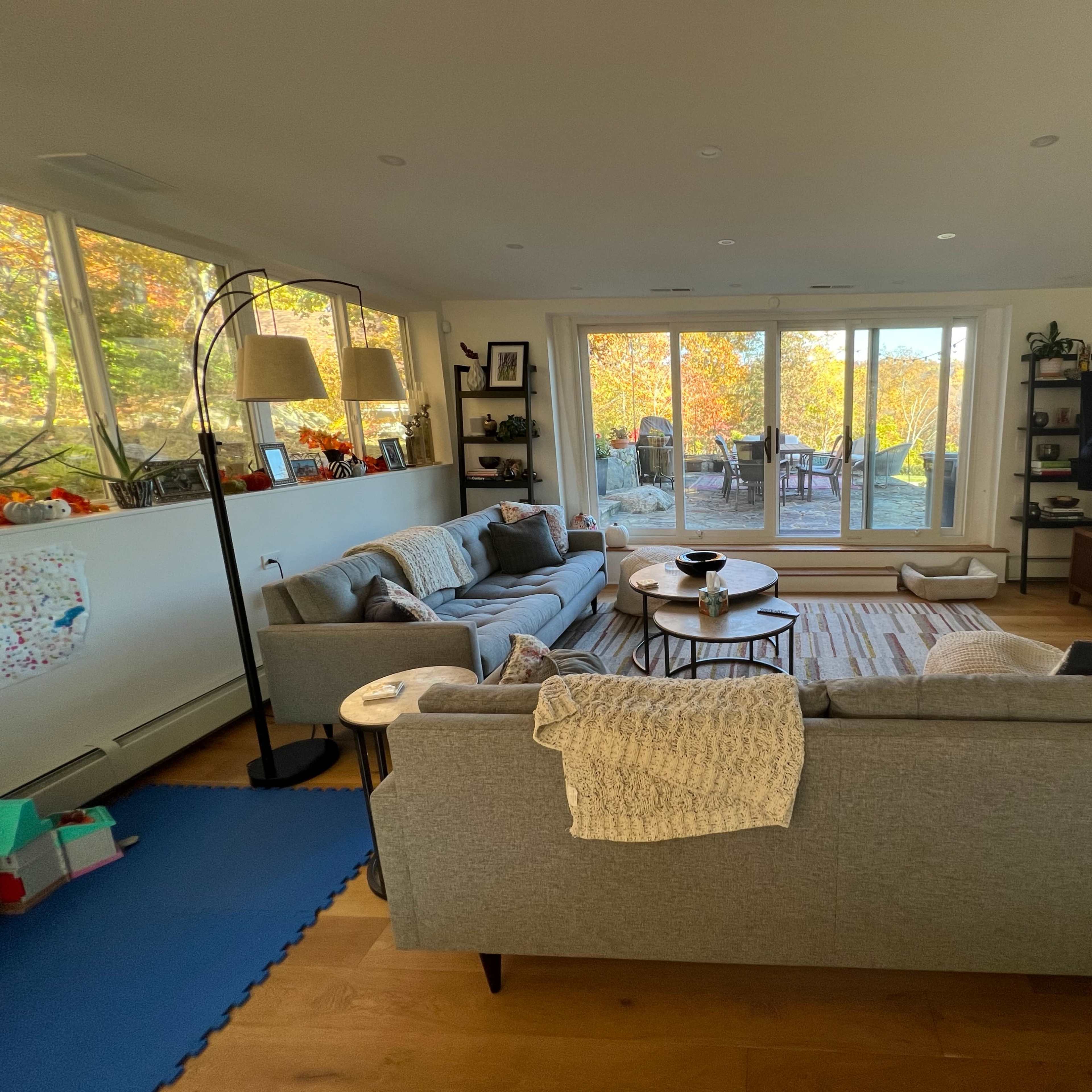 A modern living room features two gray sofas facing a large window with views of an outdoor patio and autumn foliage.