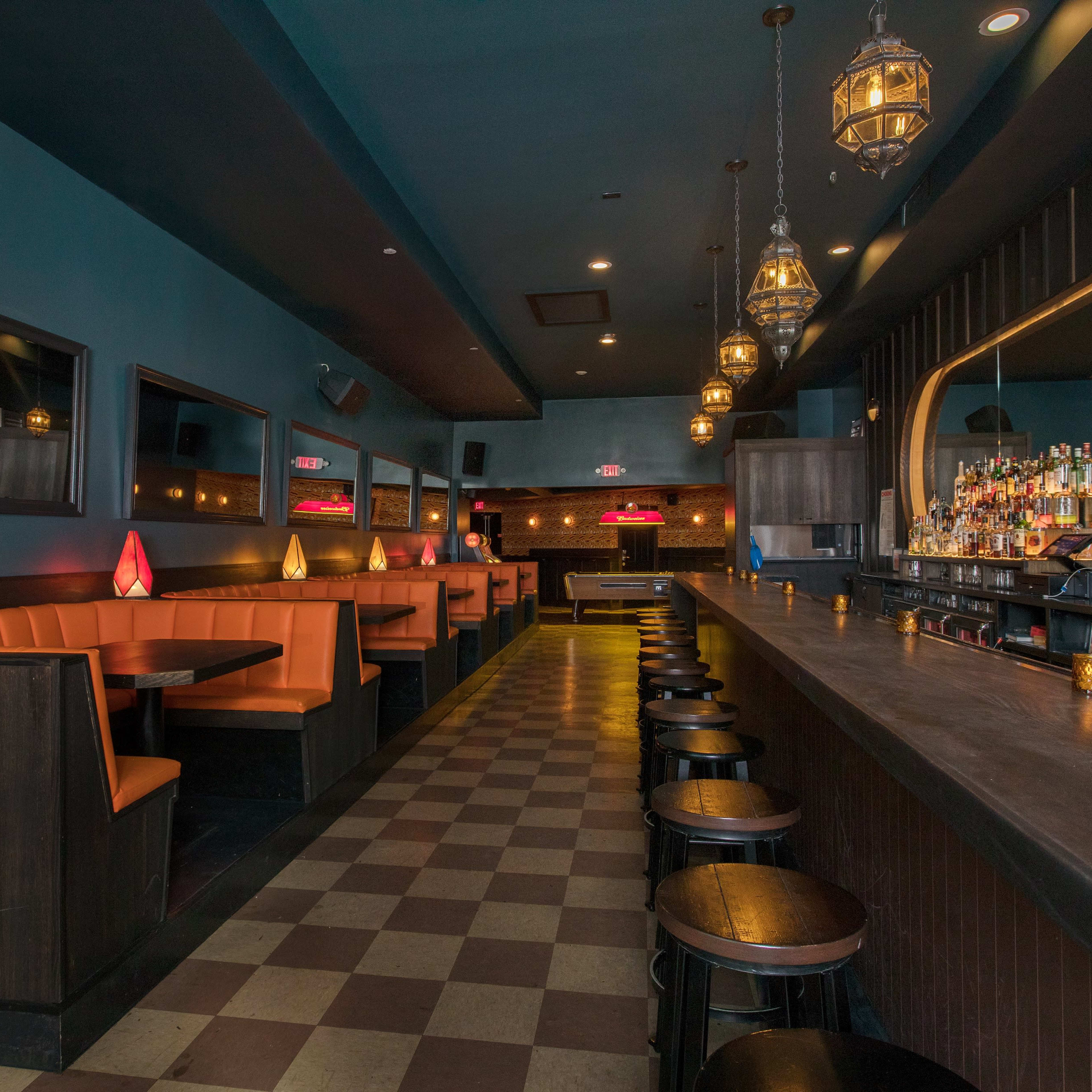 An empty bar and dining area with orange booths, a long counter with black stools, and decorative hanging lights.