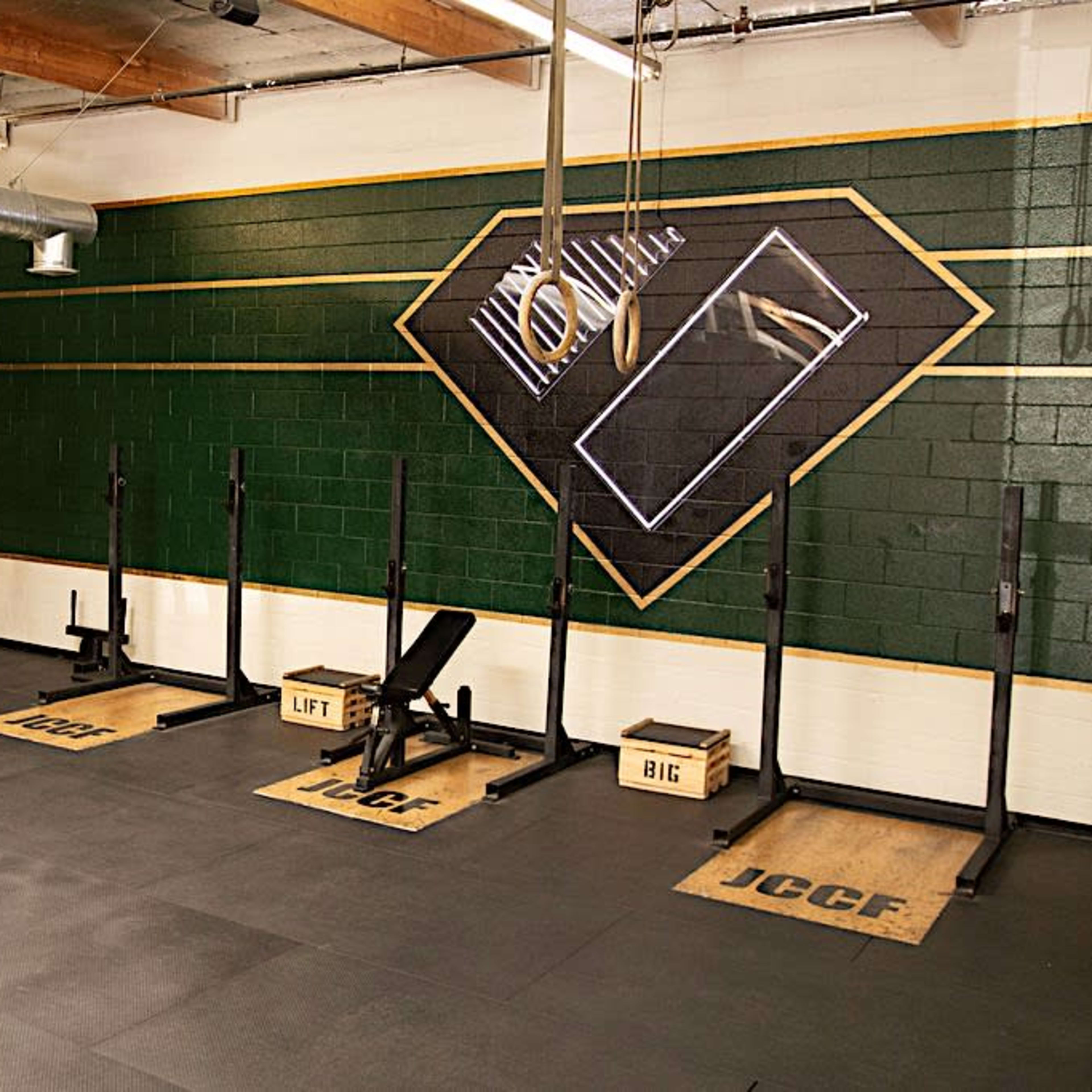 A gym with dark green walls, featuring multiple weightlifting racks and mats labeled "JCCF," "LIFT," and "BIG."