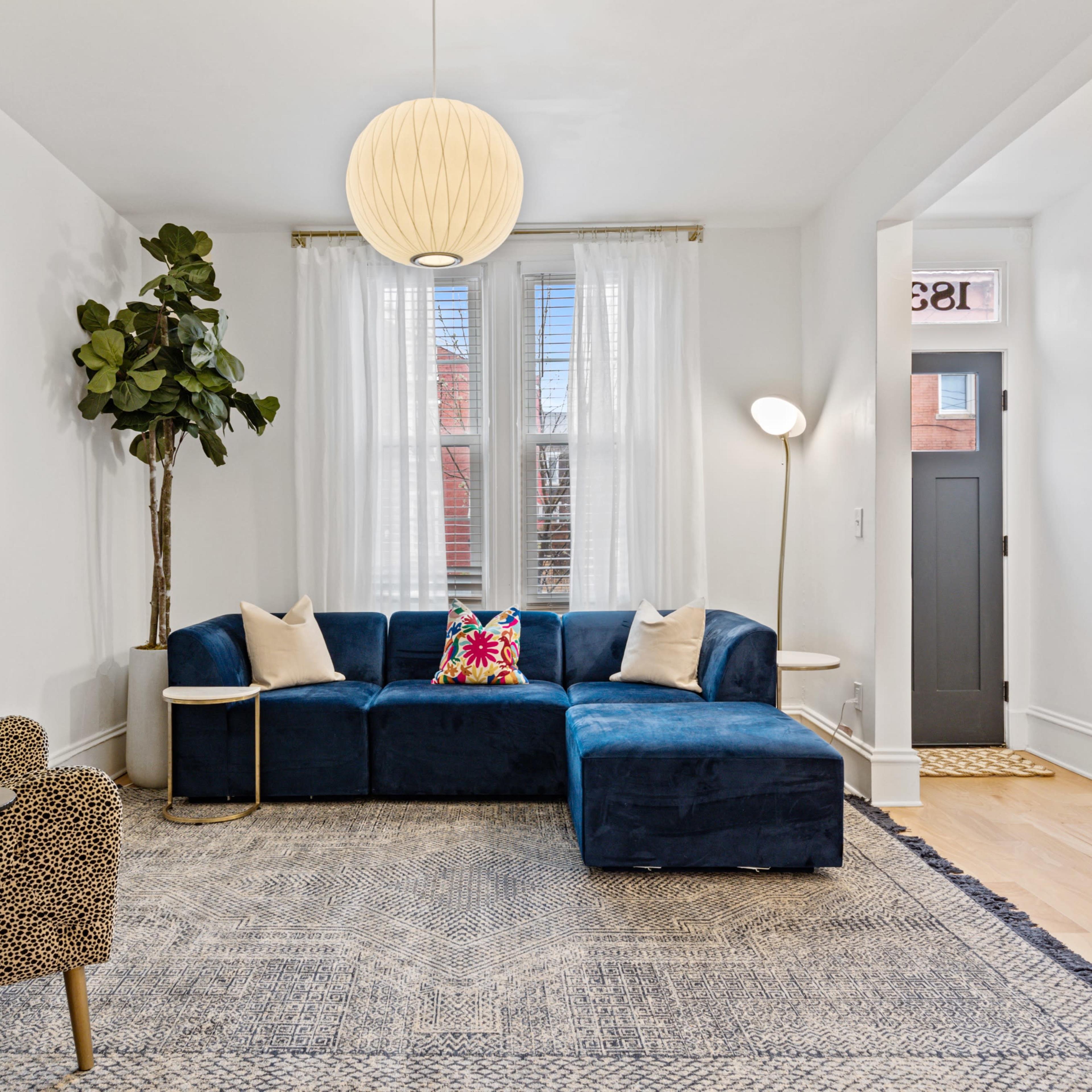 The living room features a blue sectional sofa, a patterned chair, a floor plant, and a round pendant light, complemented by a rug and framed artwork on the walls.