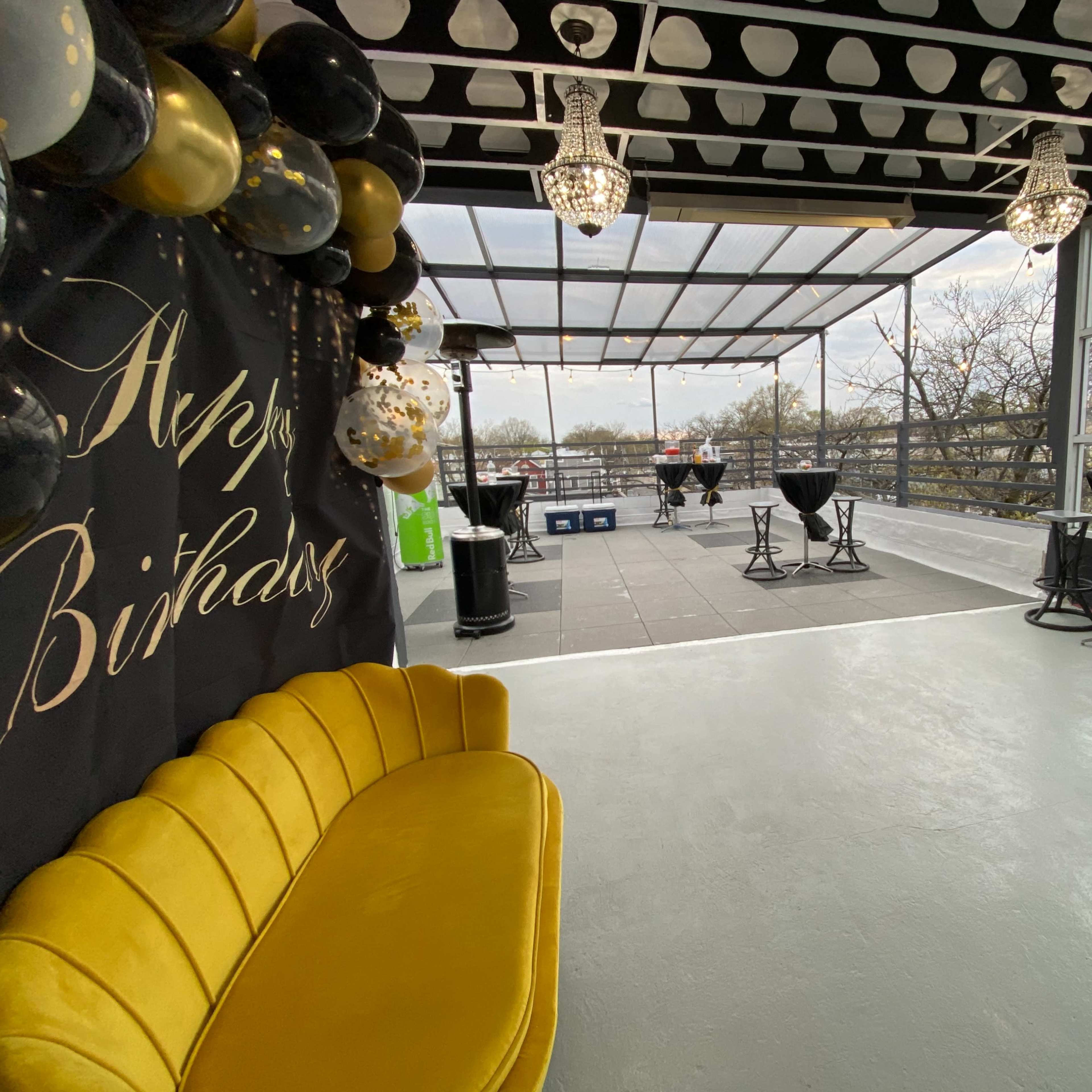 A party venue featuring a yellow couch, black and gold decorations, and a rooftop area with tables and chairs overlooking a cityscape.
