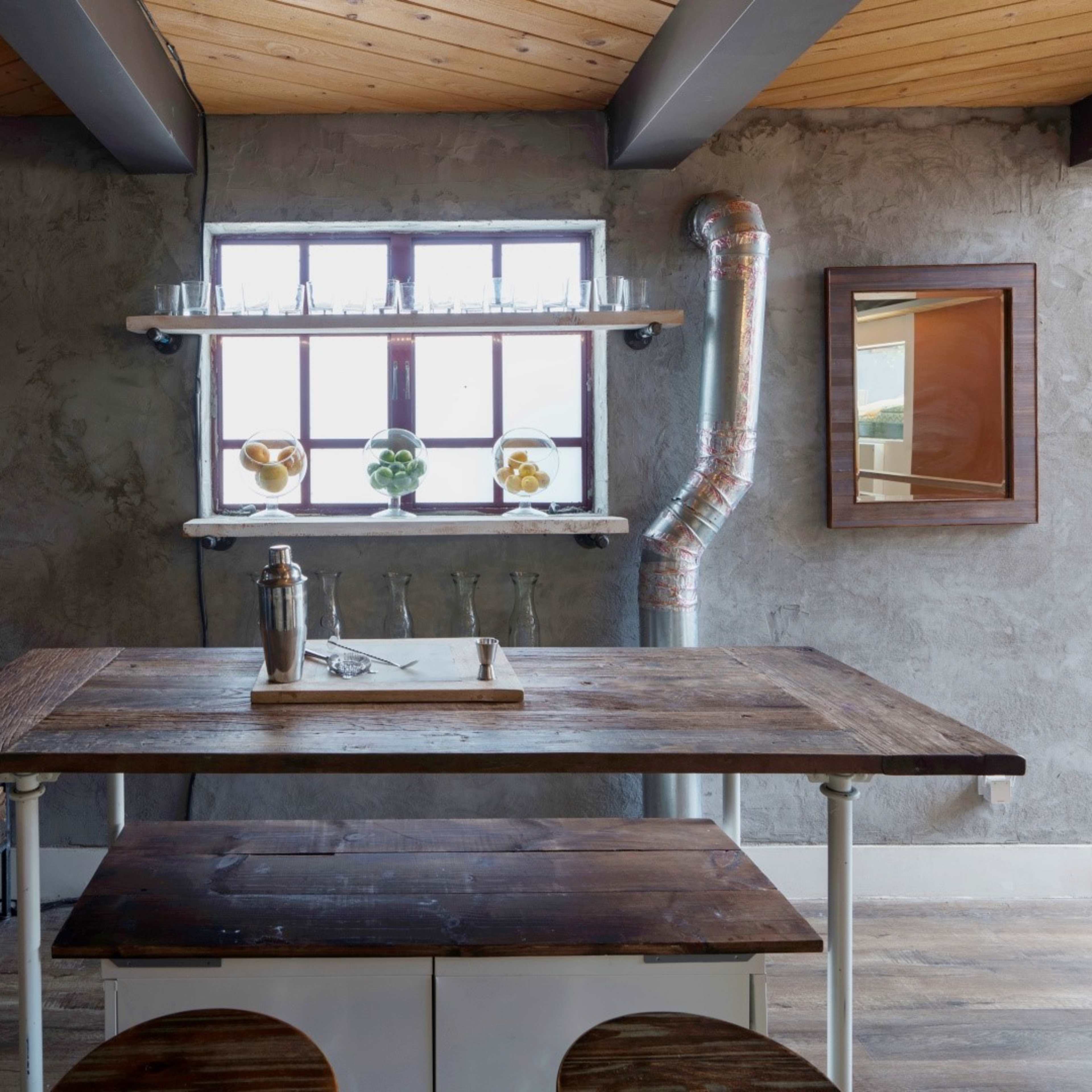 A rustic interior with a wooden table, a bench, a window featuring decorative items, and exposed pipes on a textured wall.
