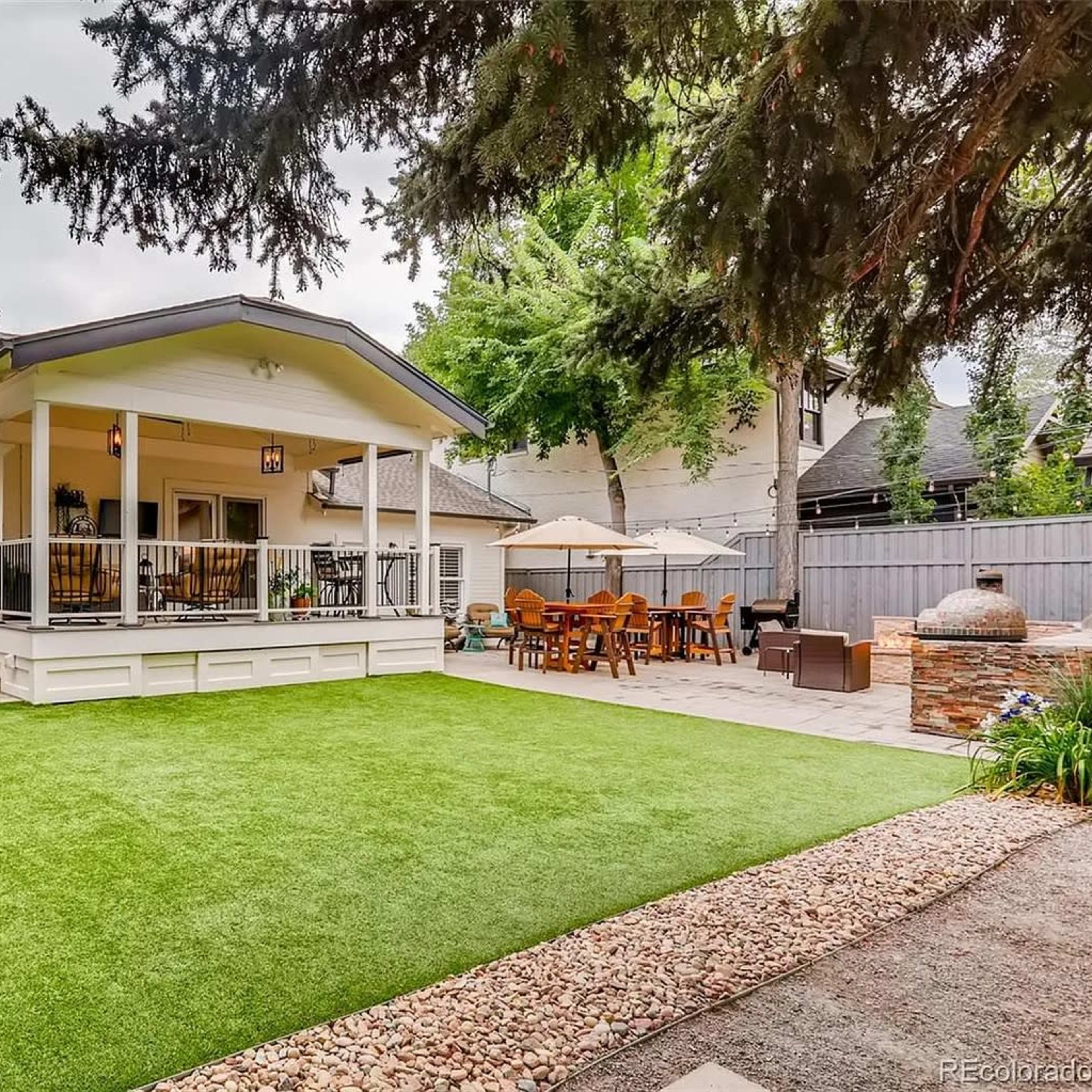 A backyard with a putting green, a covered patio area with dining furniture, and landscaping that includes trees and decorative rocks.