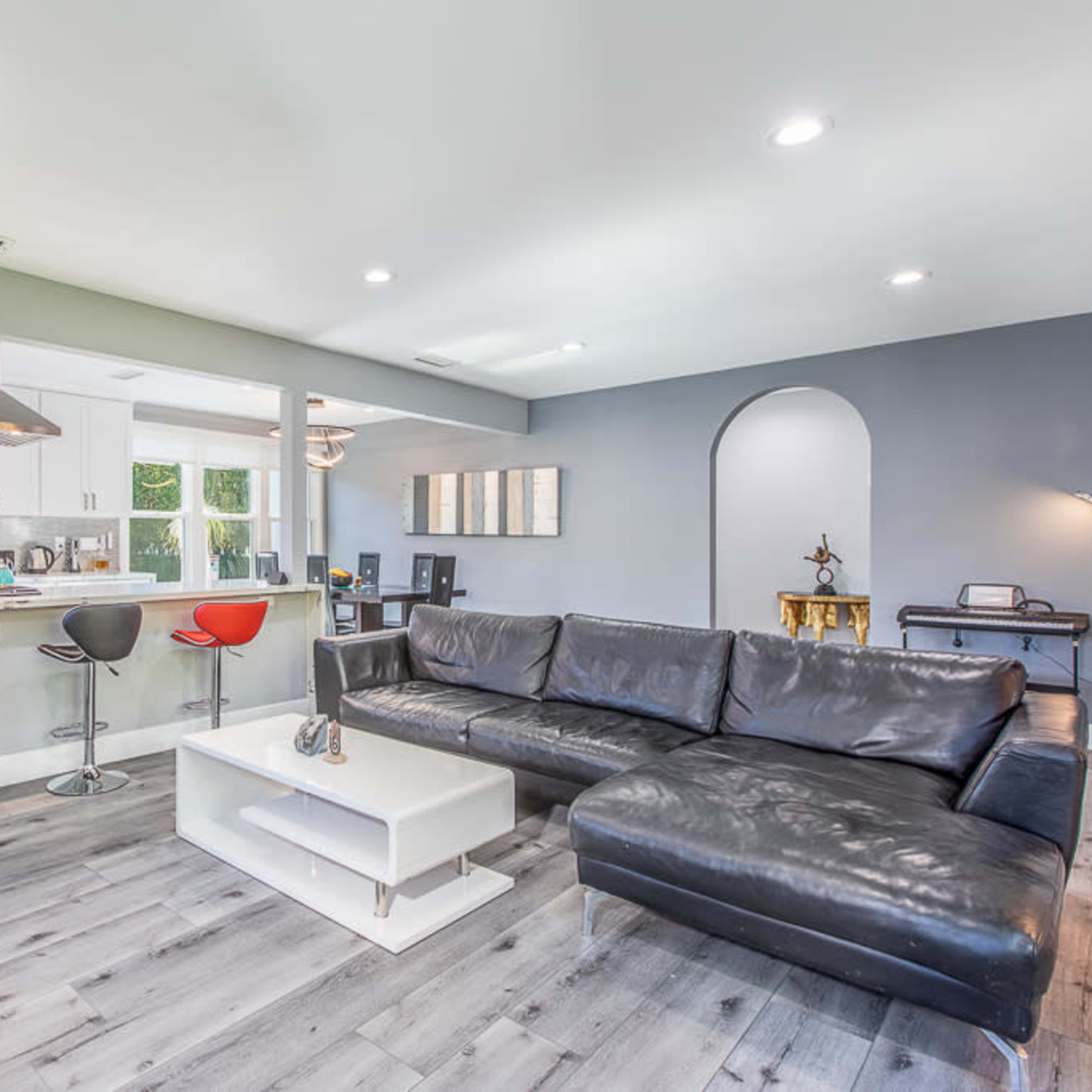 A modern living room with a black sectional sofa, a white coffee table, and a kitchen area featuring bar stools and modern appliances.