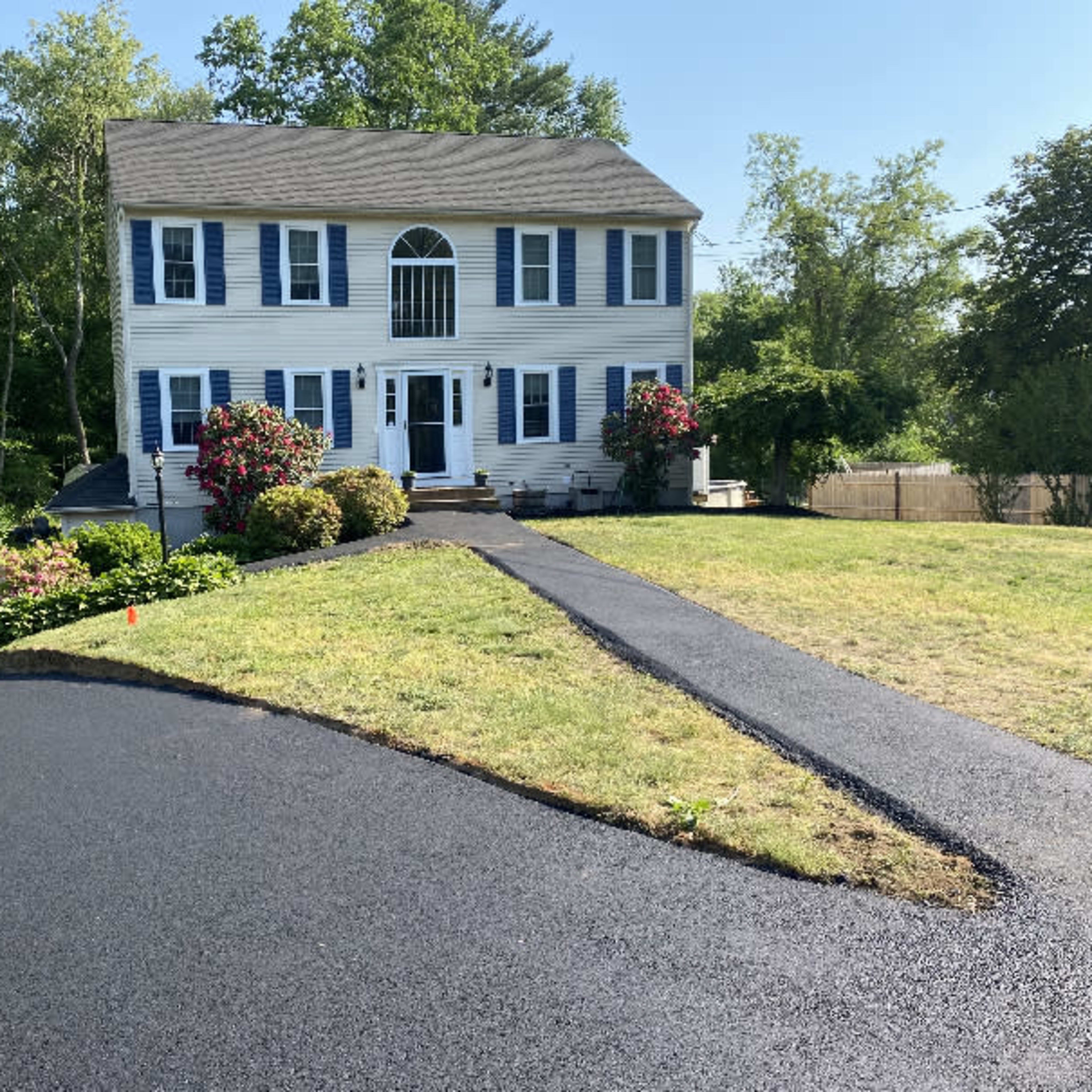 A two-story blue and white house with a front porch and landscaped yard sits on a newly asphalted driveway.