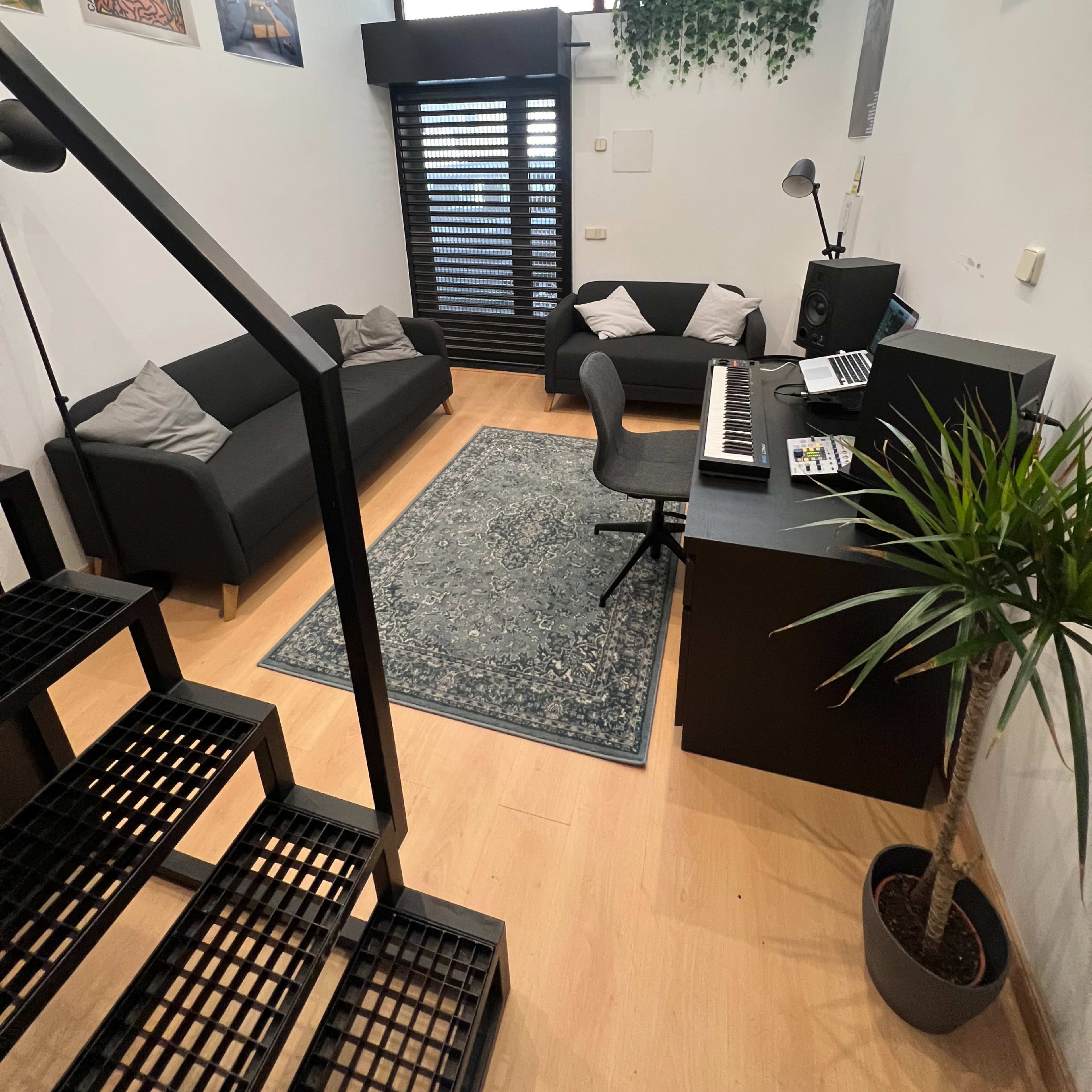 A modern room with a staircase, two black sofas, a desk with a music keyboard, and a decorative plant.