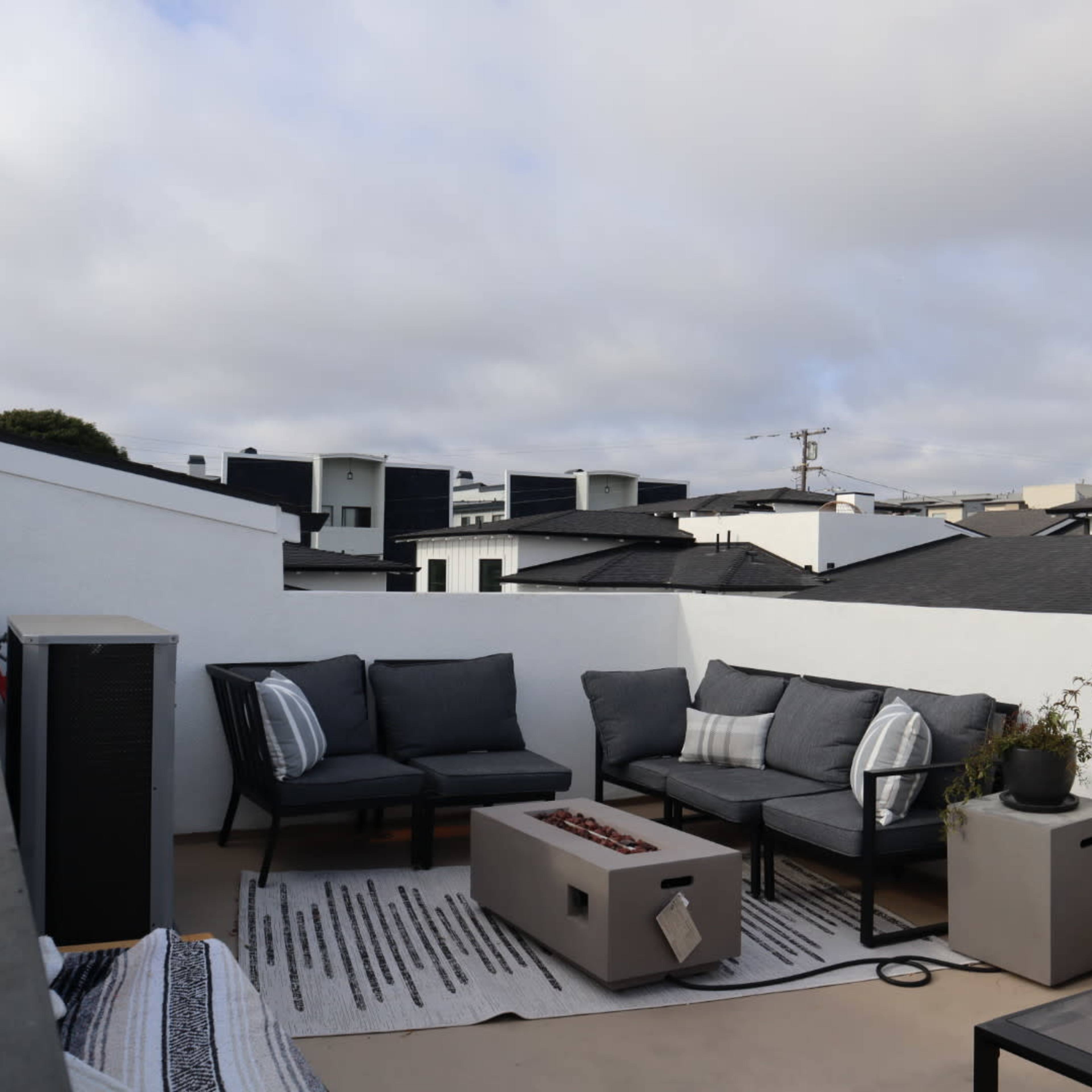 A rooftop seating area with gray outdoor furniture, a small fire pit, and a potted plant, surrounded by low white walls and rooftops of nearby buildings.
