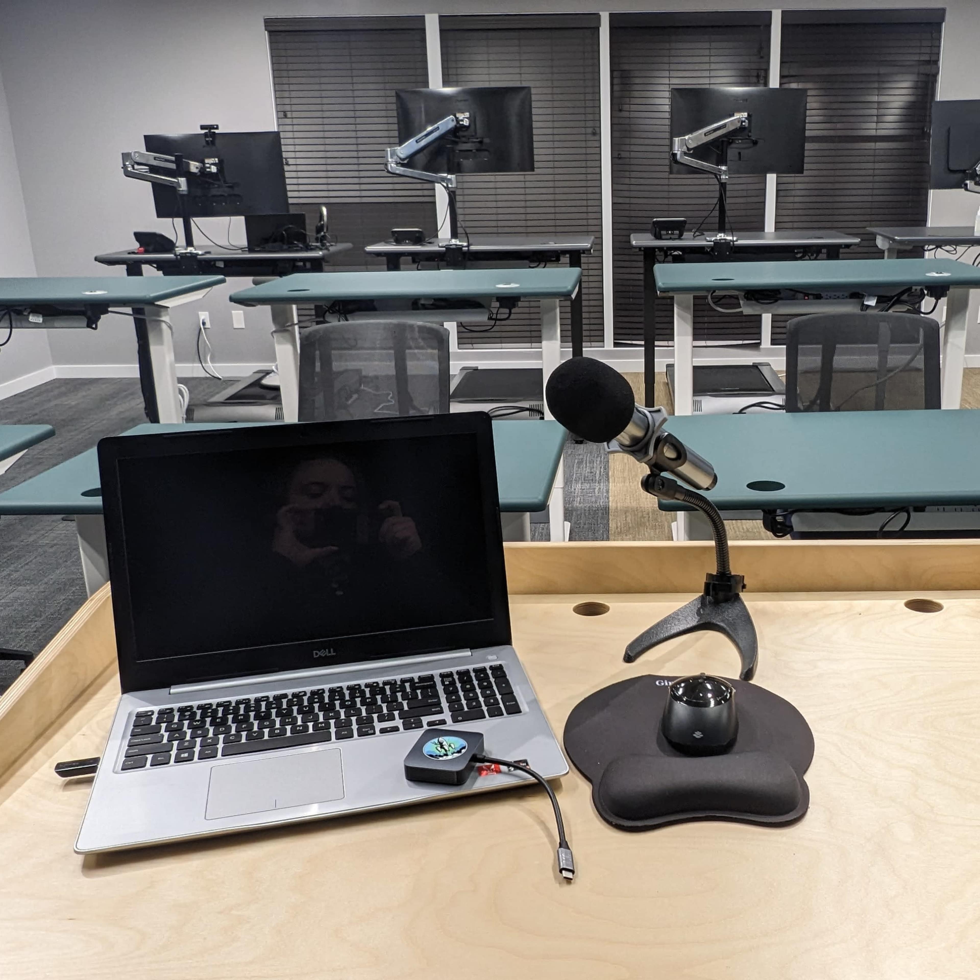 A desktop setup with a laptop and microphone is positioned at the front of a classroom filled with adjustable desks and computer monitors.