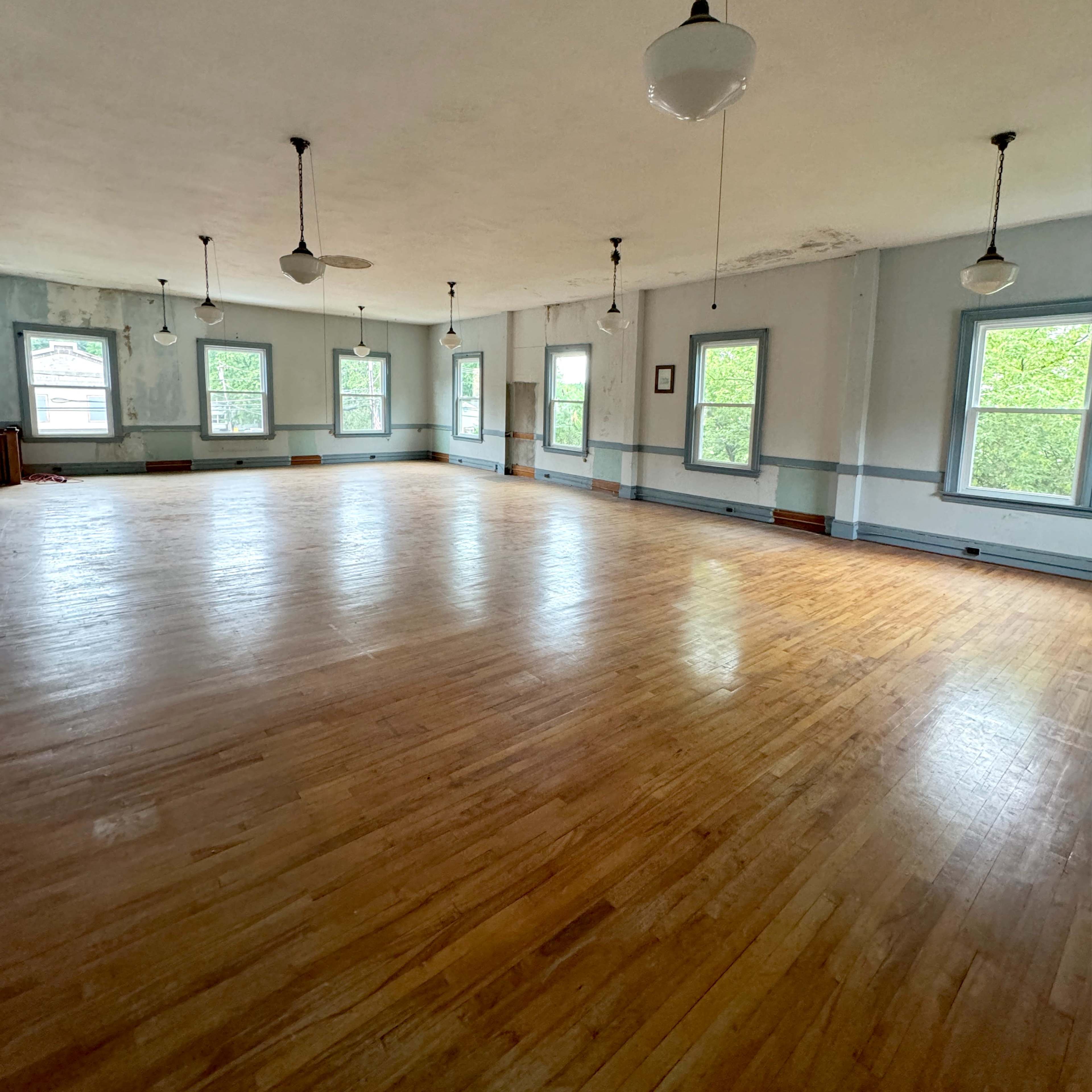 A spacious, empty room with hardwood floors and multiple windows, illuminated by hanging light fixtures.