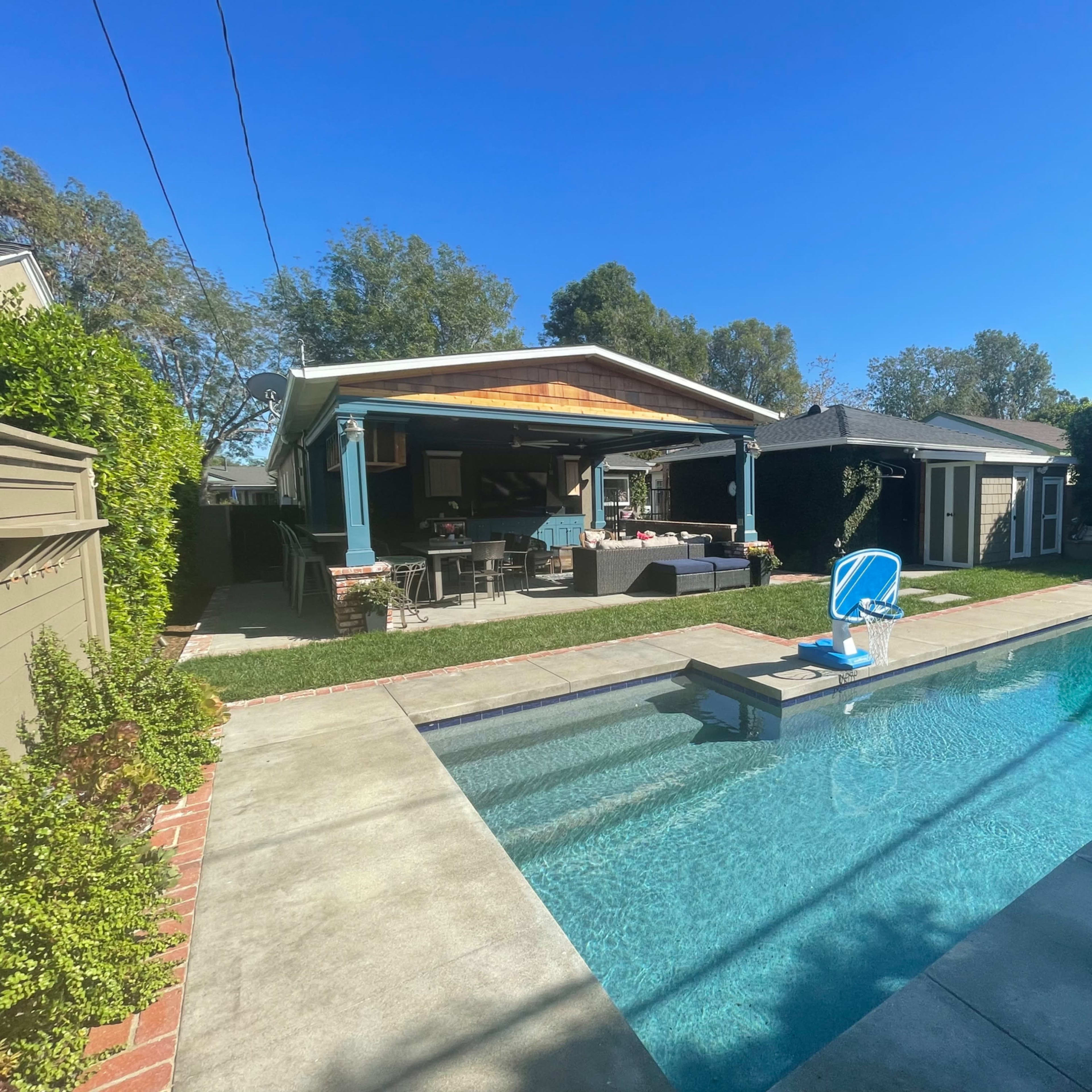 A backyard with a swimming pool surrounded by concrete, a grassy area, and a covered patio with outdoor seating.