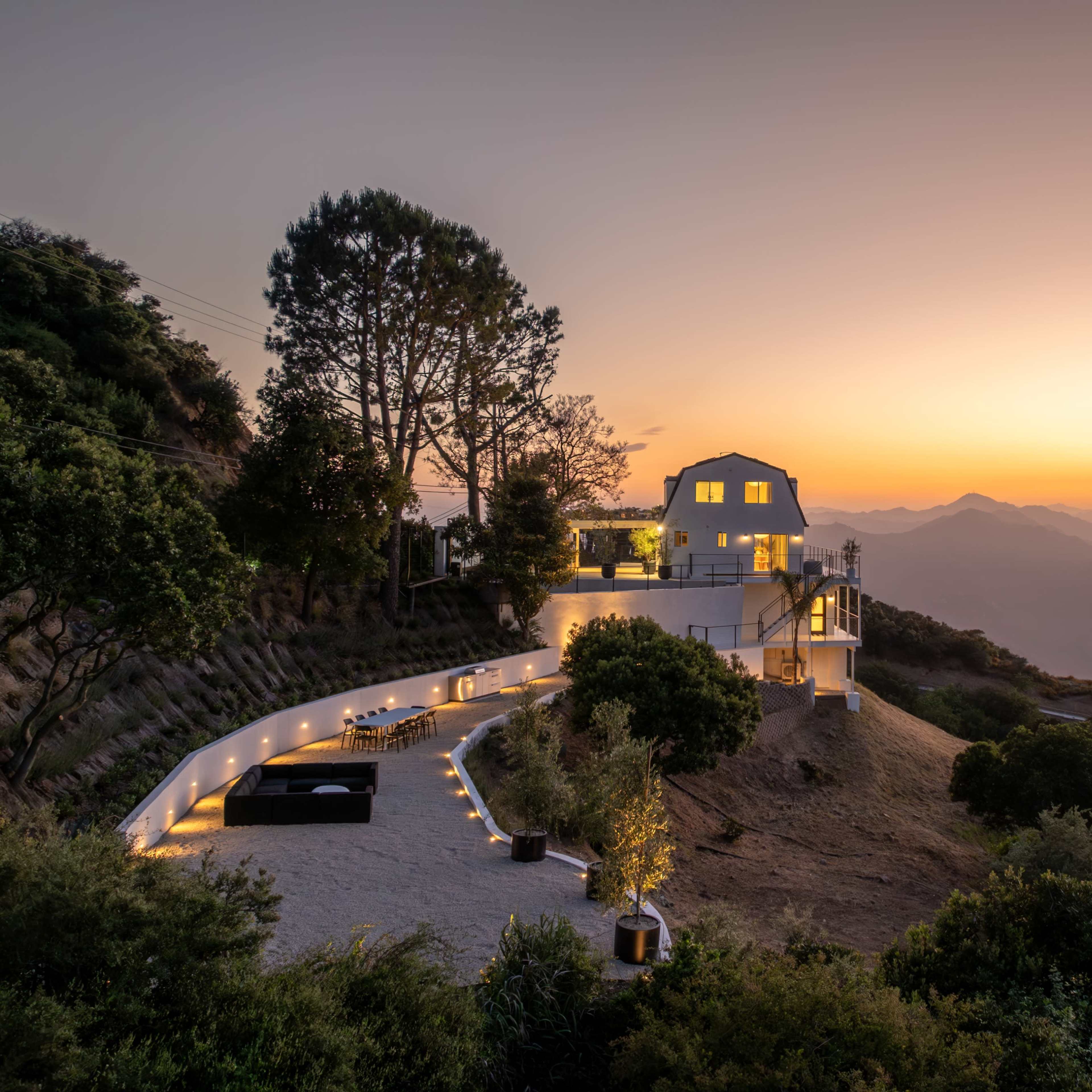 A modern white house is situated on a hillside, overlooking a mountain landscape at sunset with lights lining a paved terrace.