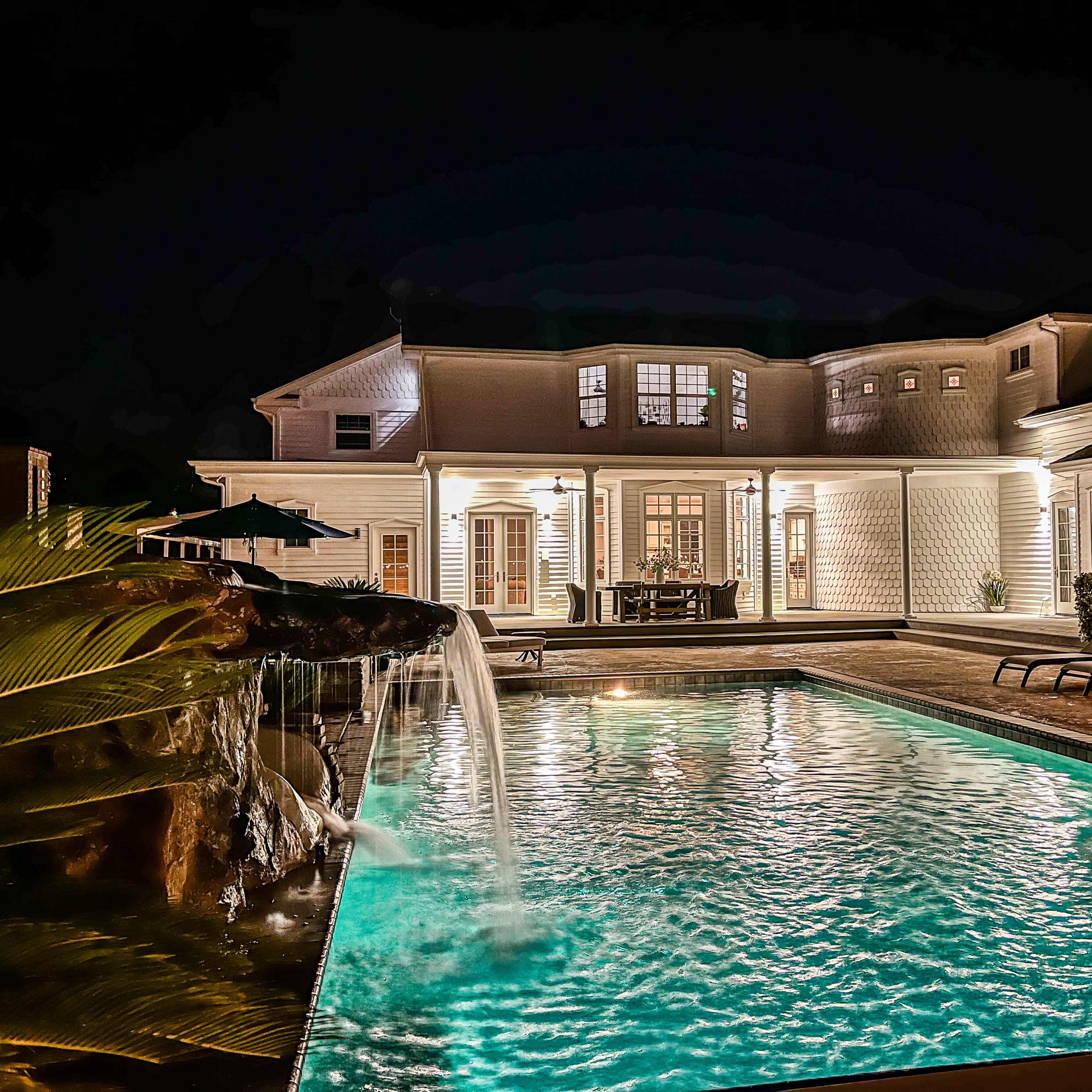A well-lit house at night with a pool and a waterfall feature, surrounded by lounge chairs and patio furniture.