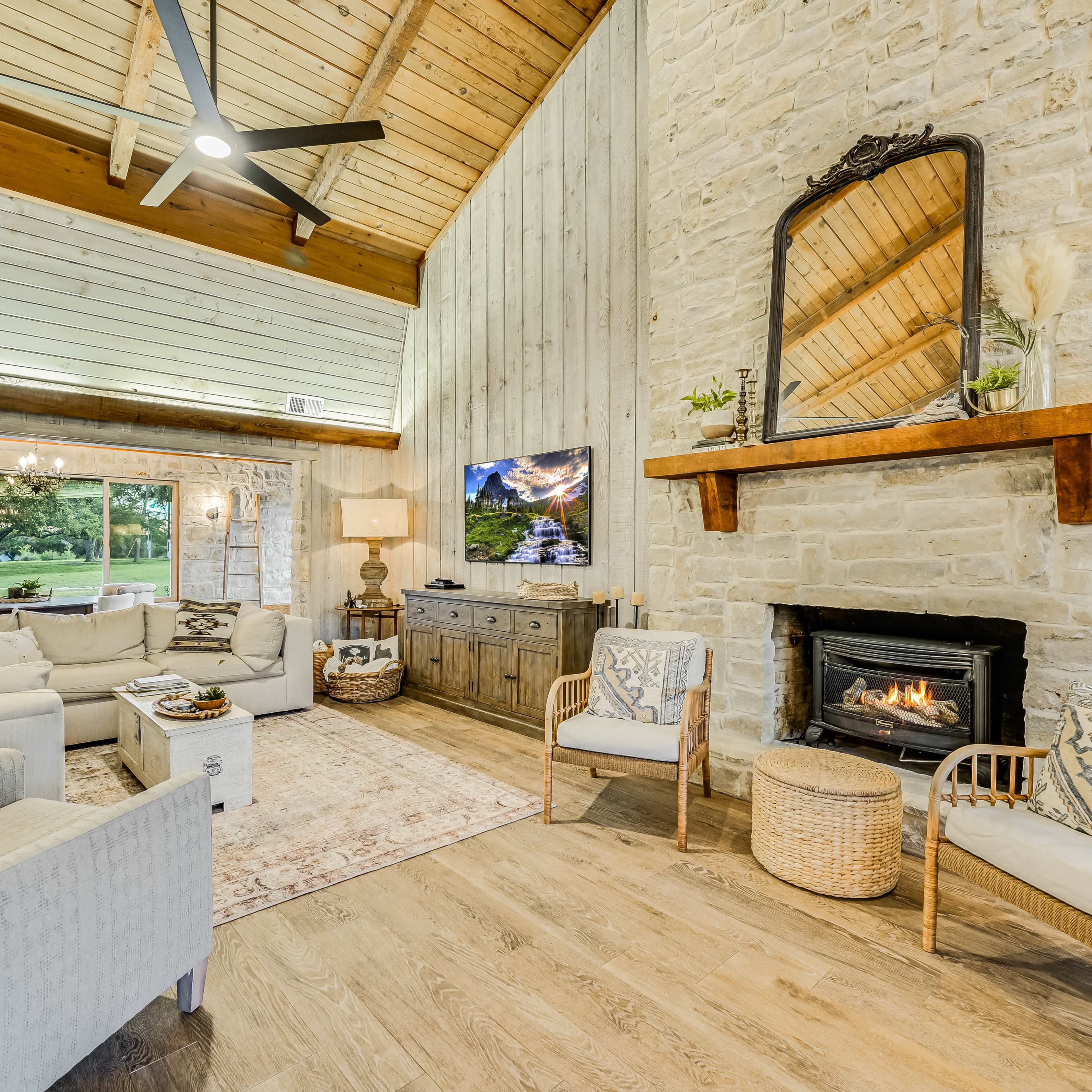 A spacious living room with a stone fireplace, wooden beams, and large windows overlooking a green outdoor area.