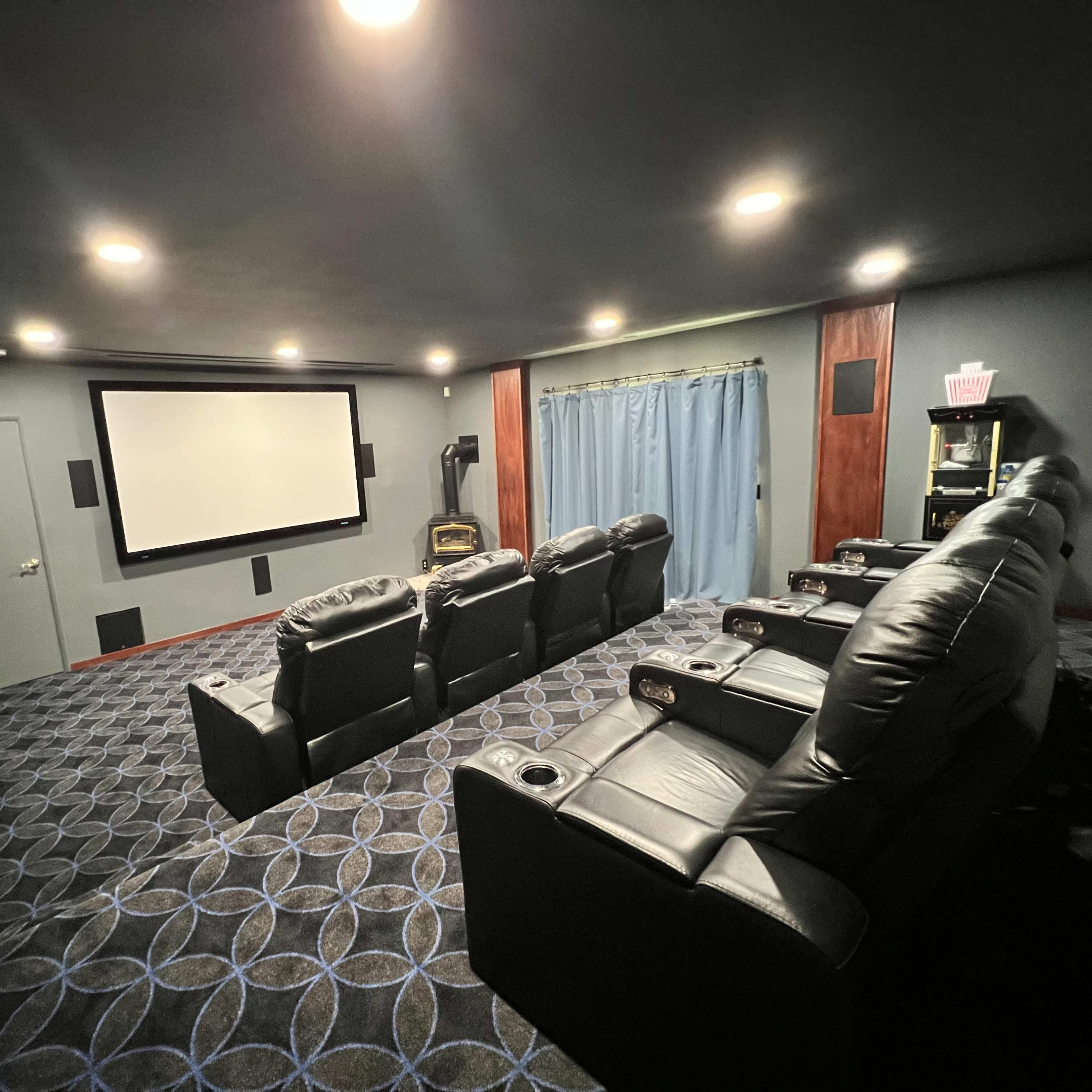 A home theater room featuring comfortable black recliner chairs arranged in front of a large projection screen, with blue curtains and a patterned carpet.