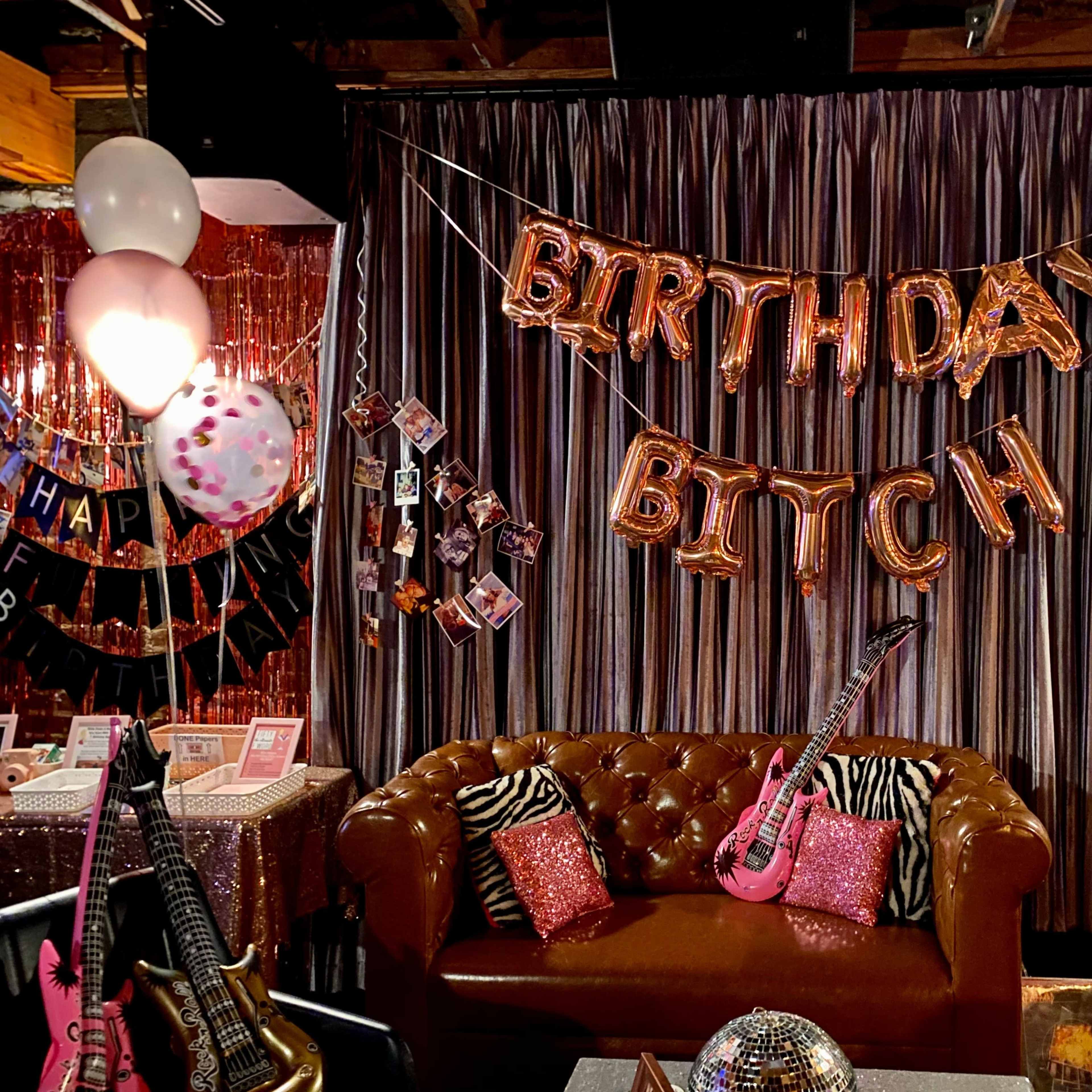 A decorated room with a "BIRTHDAY BITCH" balloon banner, seating area, and various party decorations.