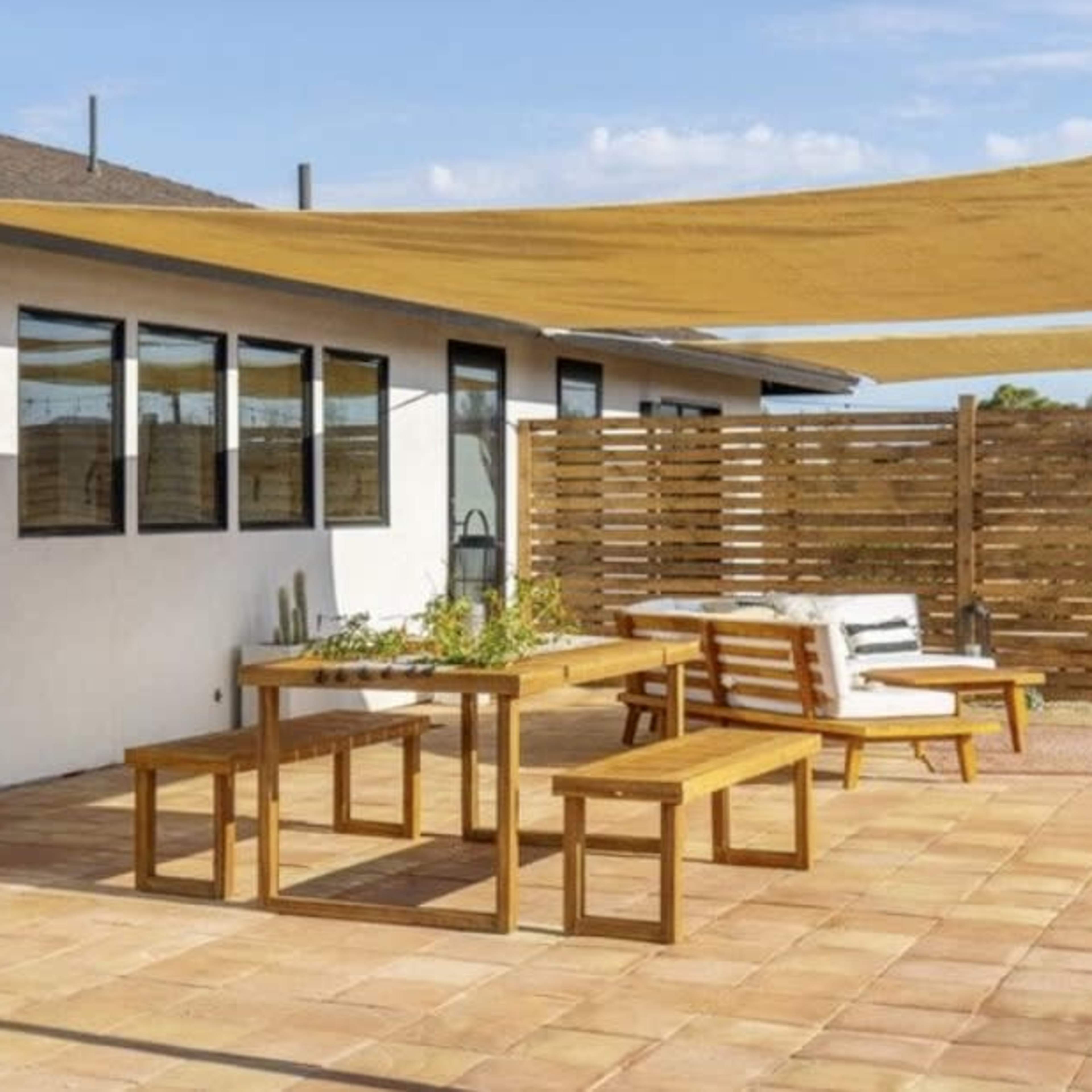 A patio area with wooden furniture, including a table and benches, surrounded by a fence and covered with a shade sail.