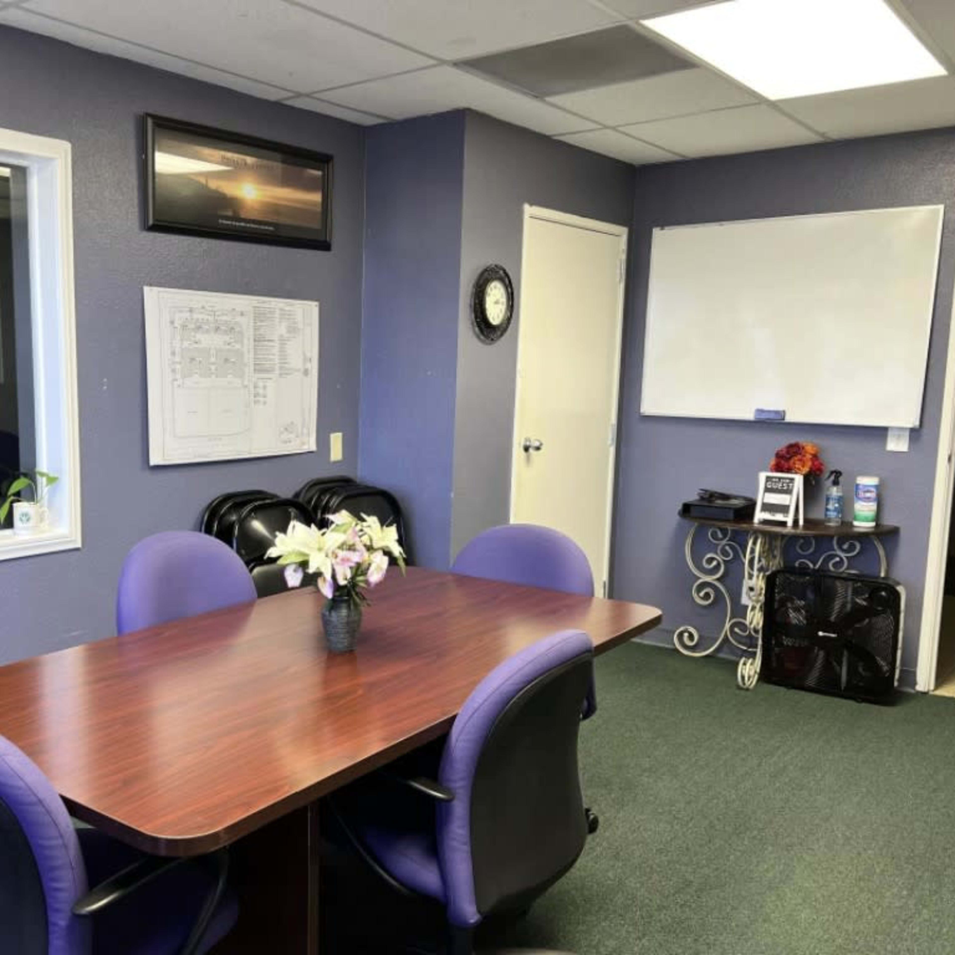 A meeting room features a brown table surrounded by black and purple chairs, with a window, a wall-mounted TV, a whiteboard, and various office supplies.