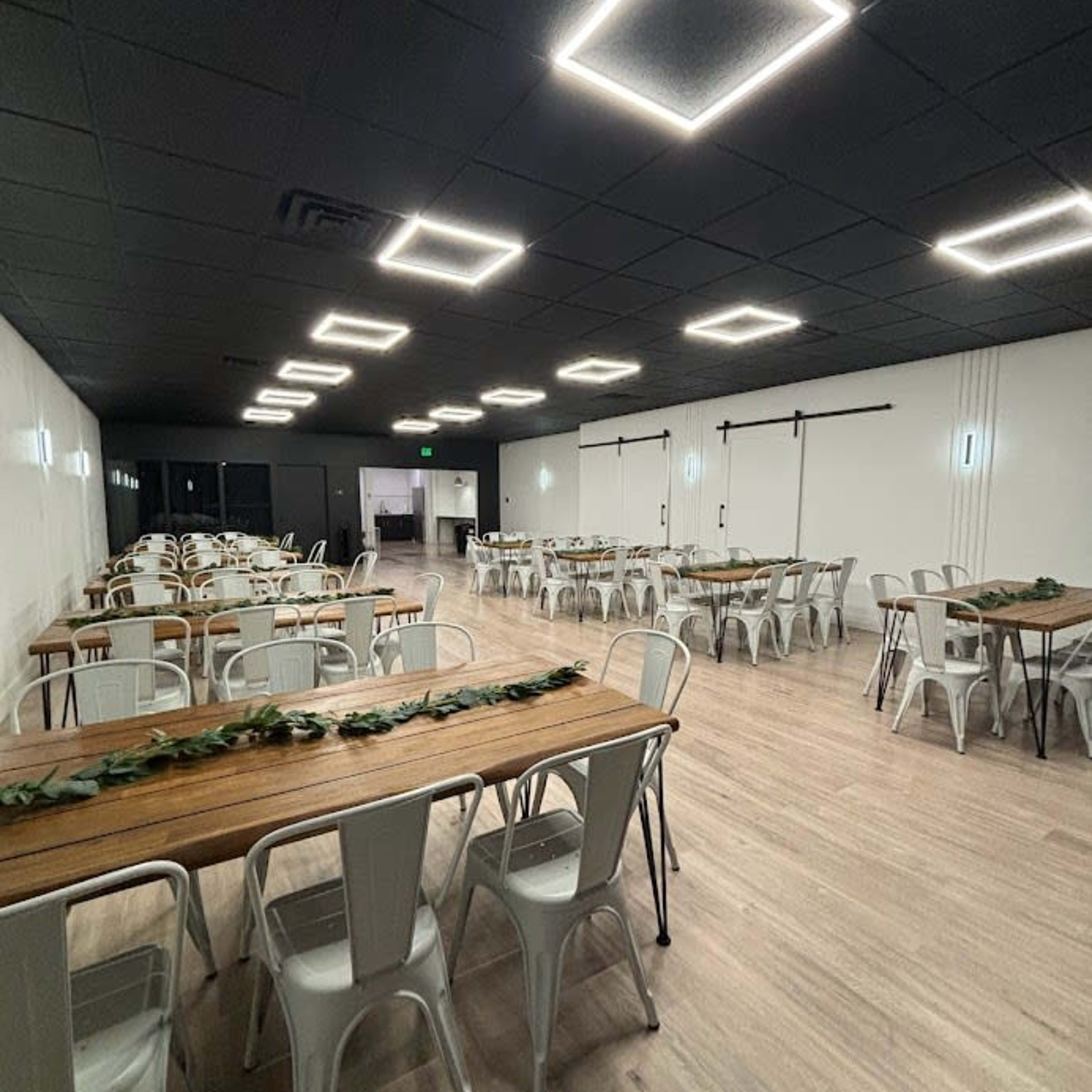 A spacious event room featuring several long wooden tables surrounded by metal chairs, illuminated by square ceiling lights.