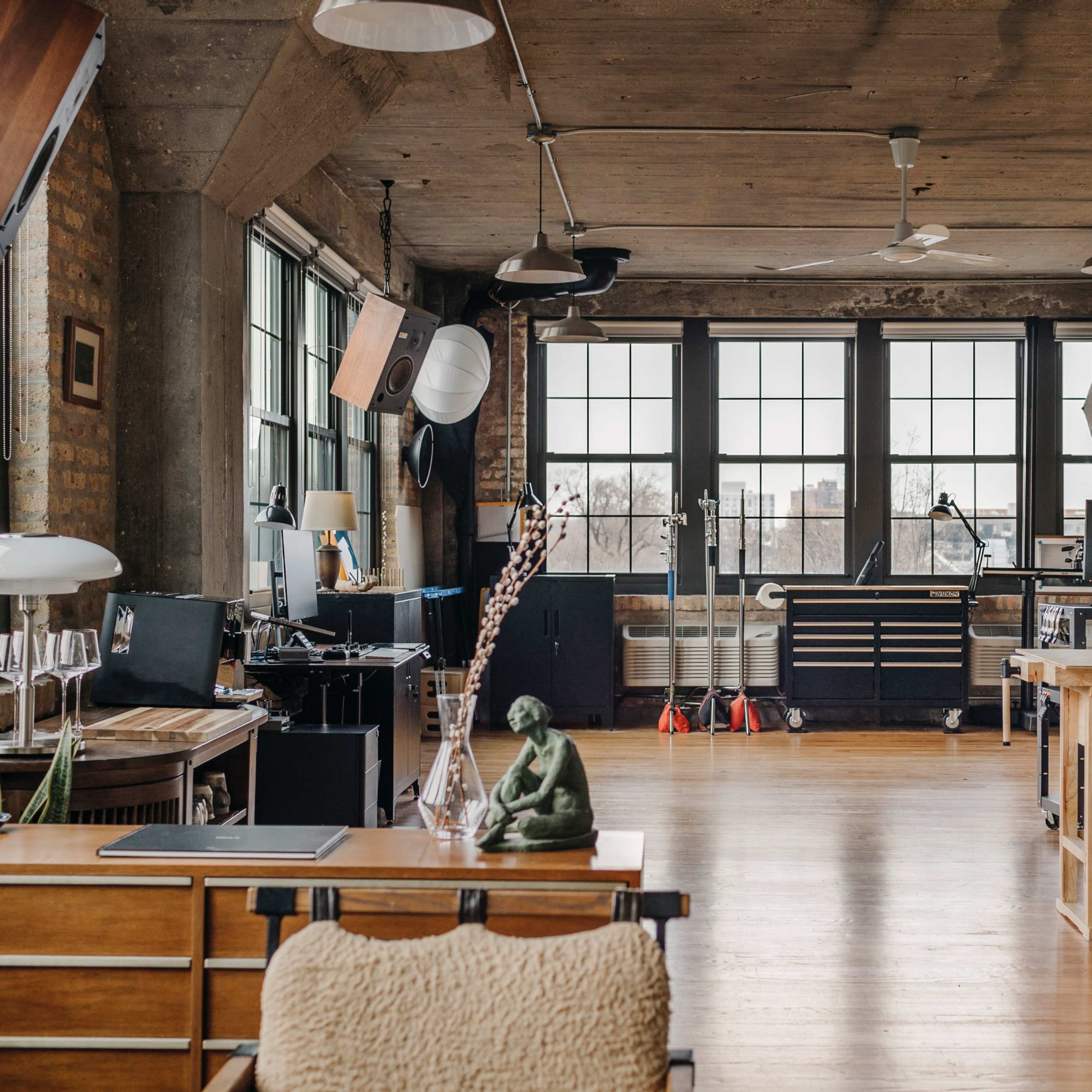 A spacious, industrial-style studio with large windows, wooden flooring, and various workstations, including a desk and a long table.