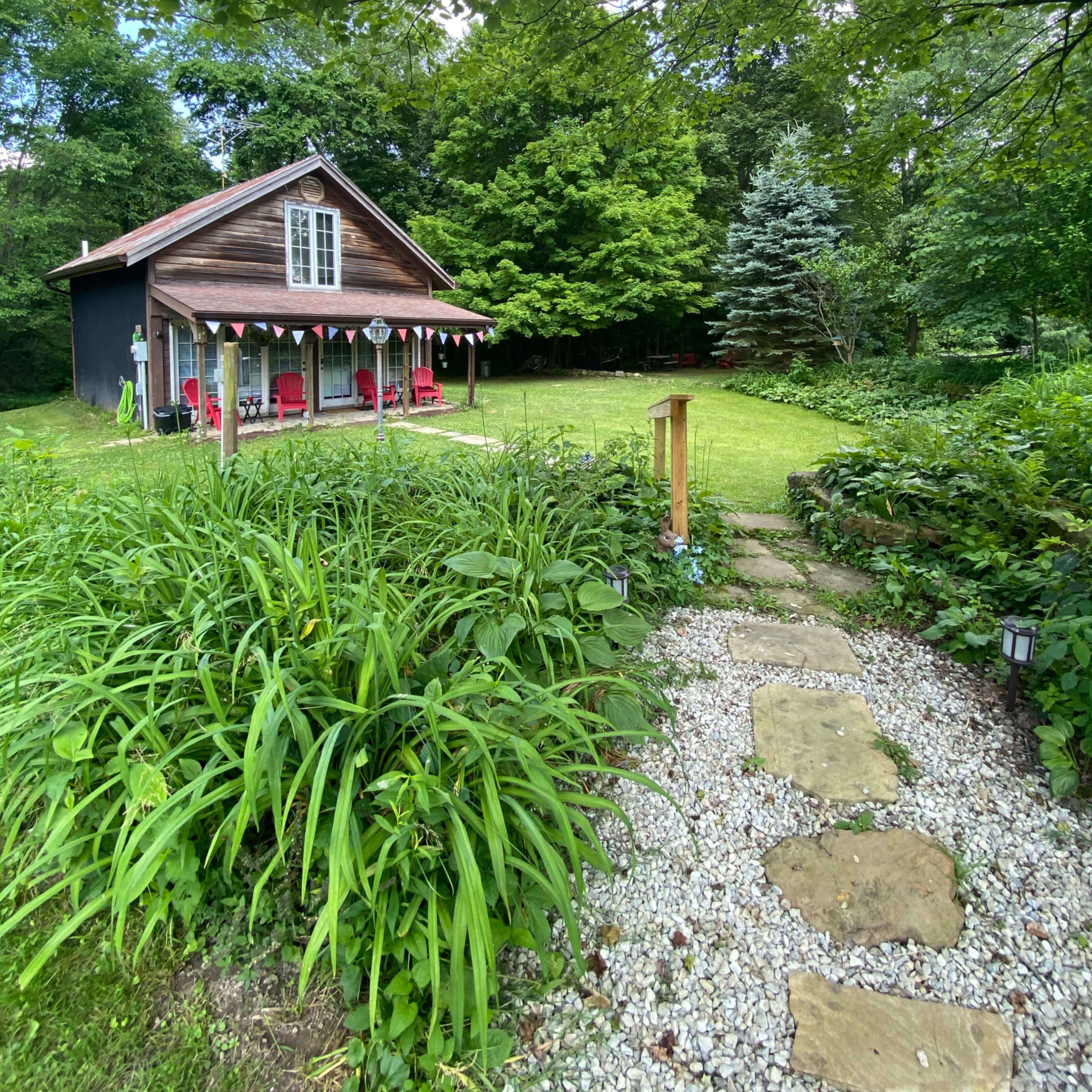 A stone pathway leads through lush greenery to a wooden house with a porch decorated with chairs and bunting.