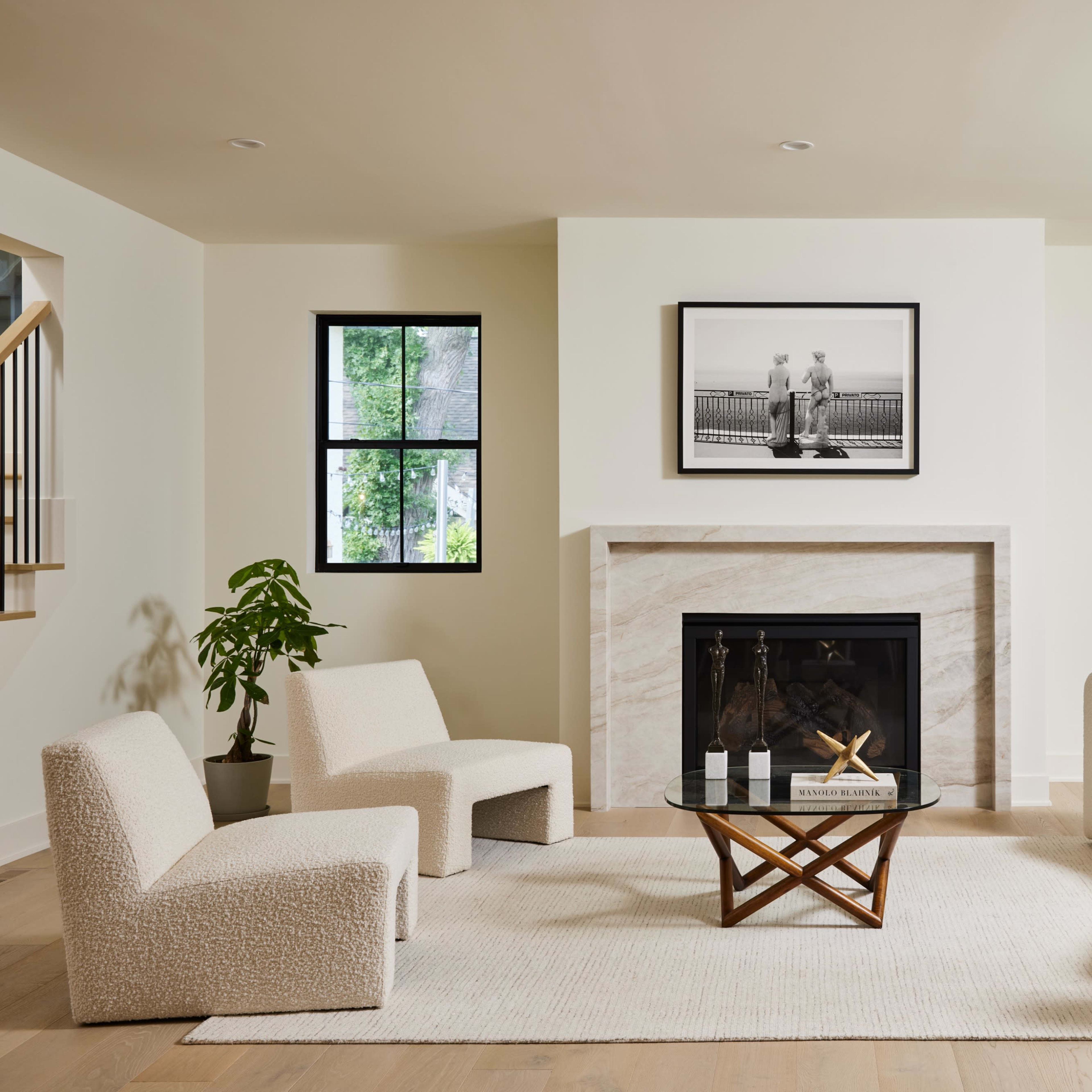 A modern living room features two upholstered chairs, a round sofa, a glass coffee table on a rug, and a fireplace with a framed black-and-white photograph above it.