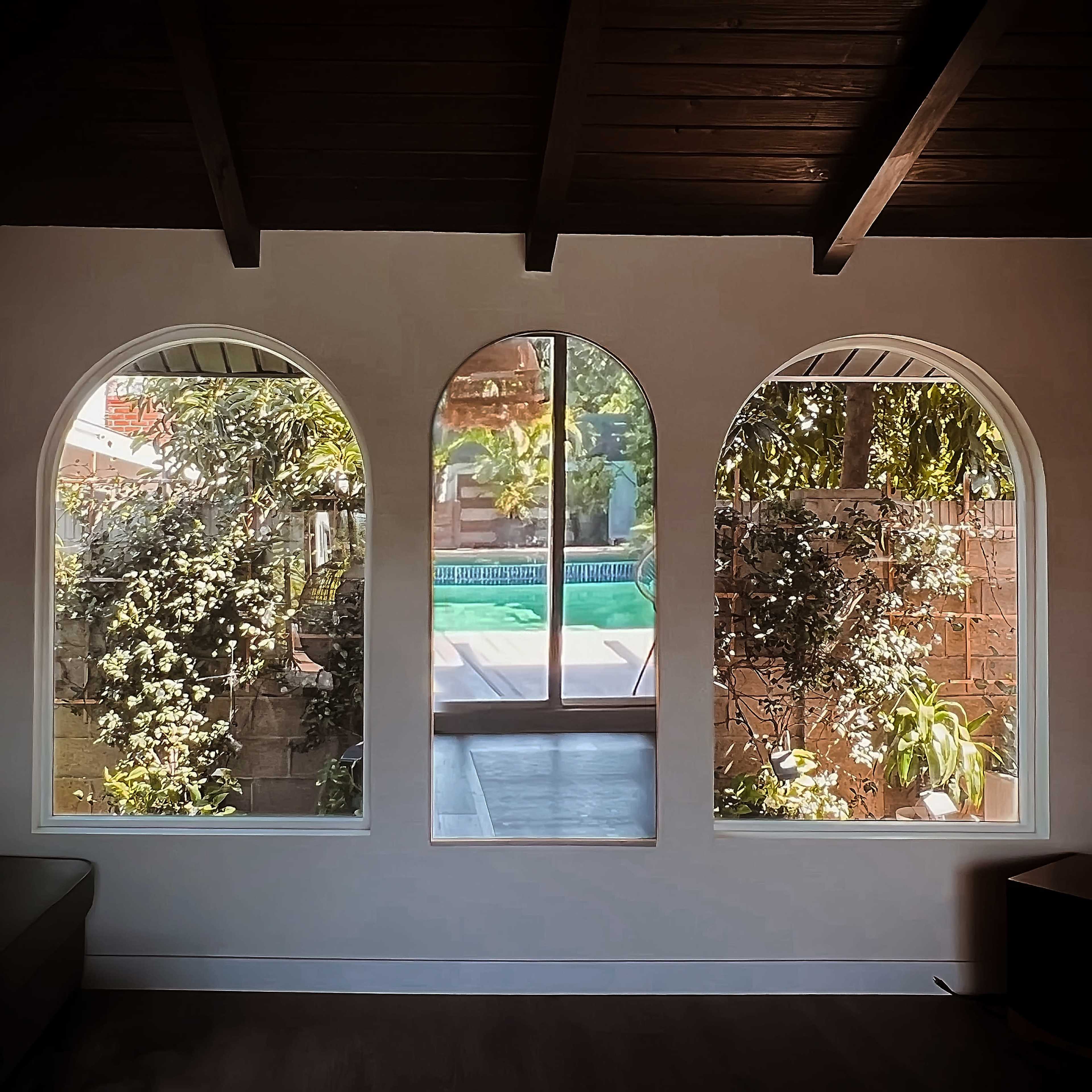 Three arched windows framed by dark wooden beams, revealing a view of a landscaped outdoor area with a swimming pool and greenery.