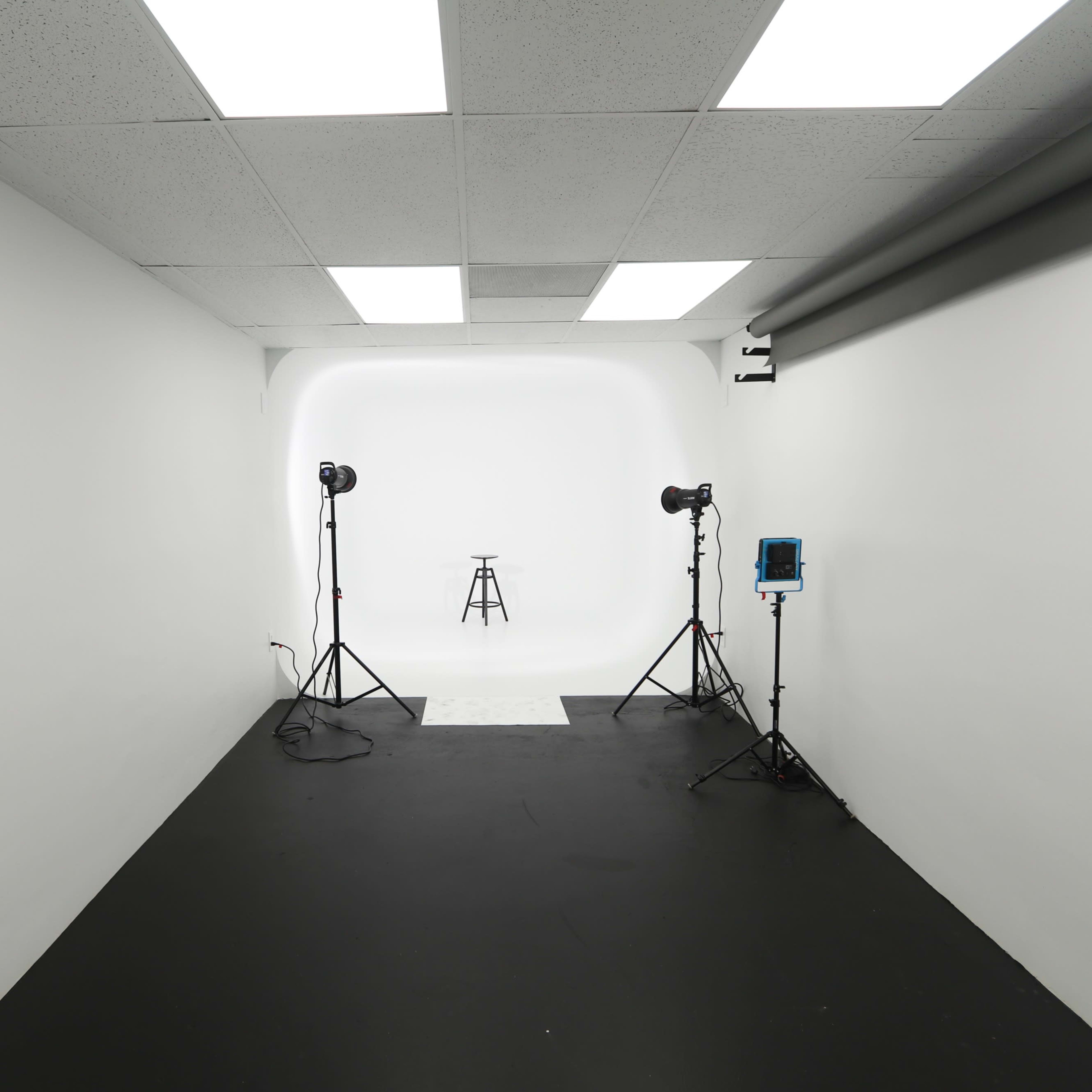 A clean, empty photography studio features a black floor, white walls, and a solitary stool illuminated by softbox lights on tripods.